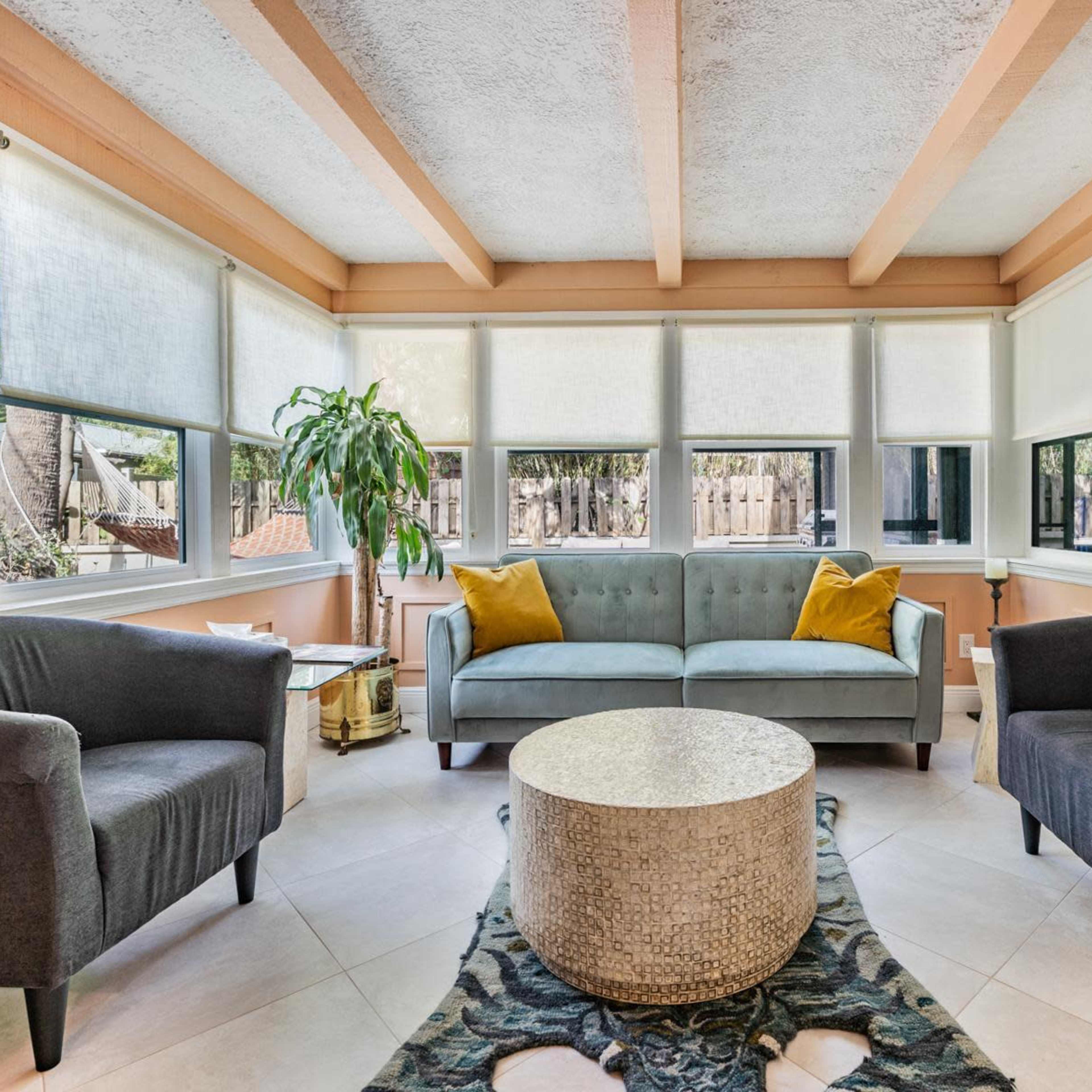 A sunlit sitting area features a gray sofa with yellow cushions, two matching armchairs, and a round stone coffee table, surrounded by large windows.