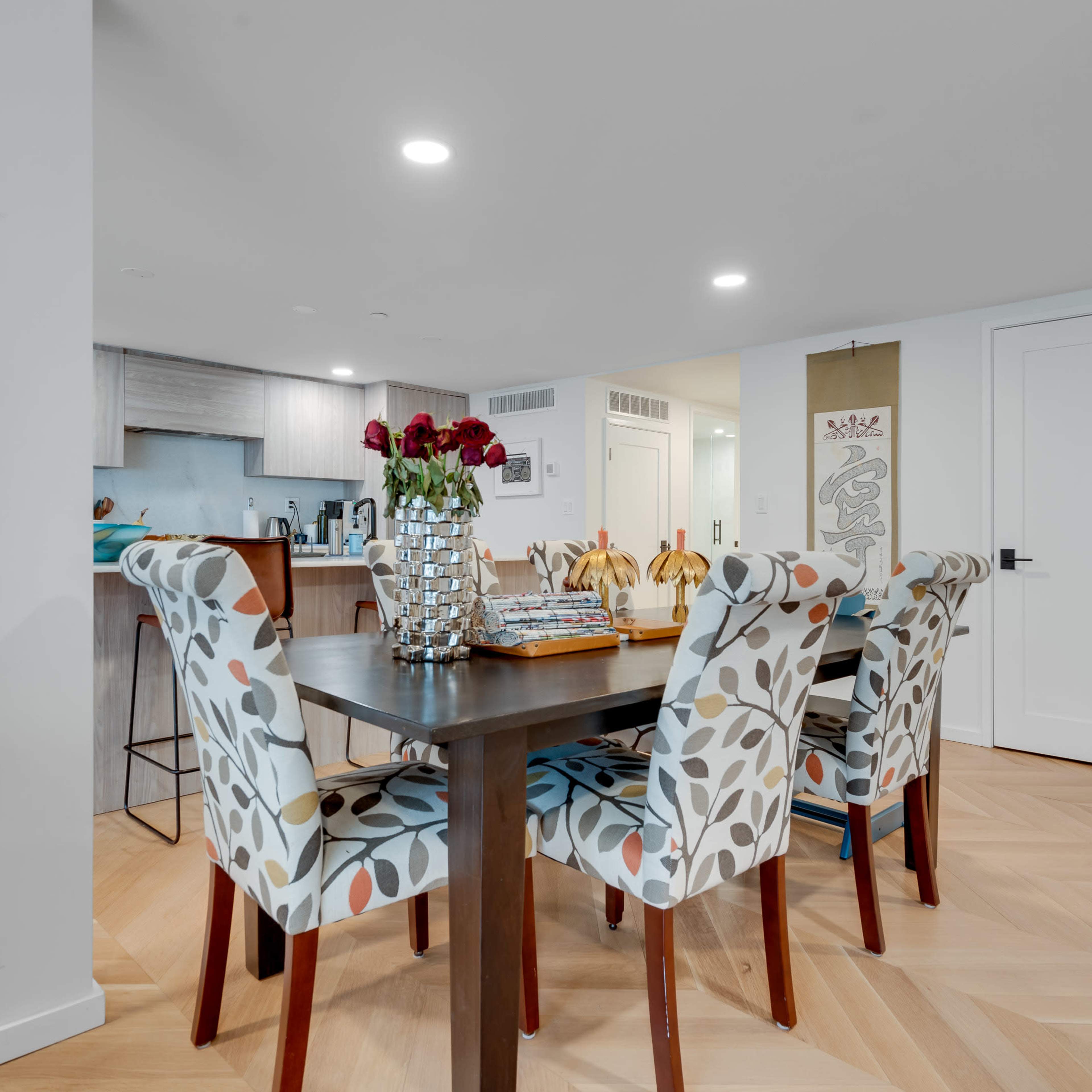 A dining area features a table surrounded by four patterned chairs with a vase of roses on top.