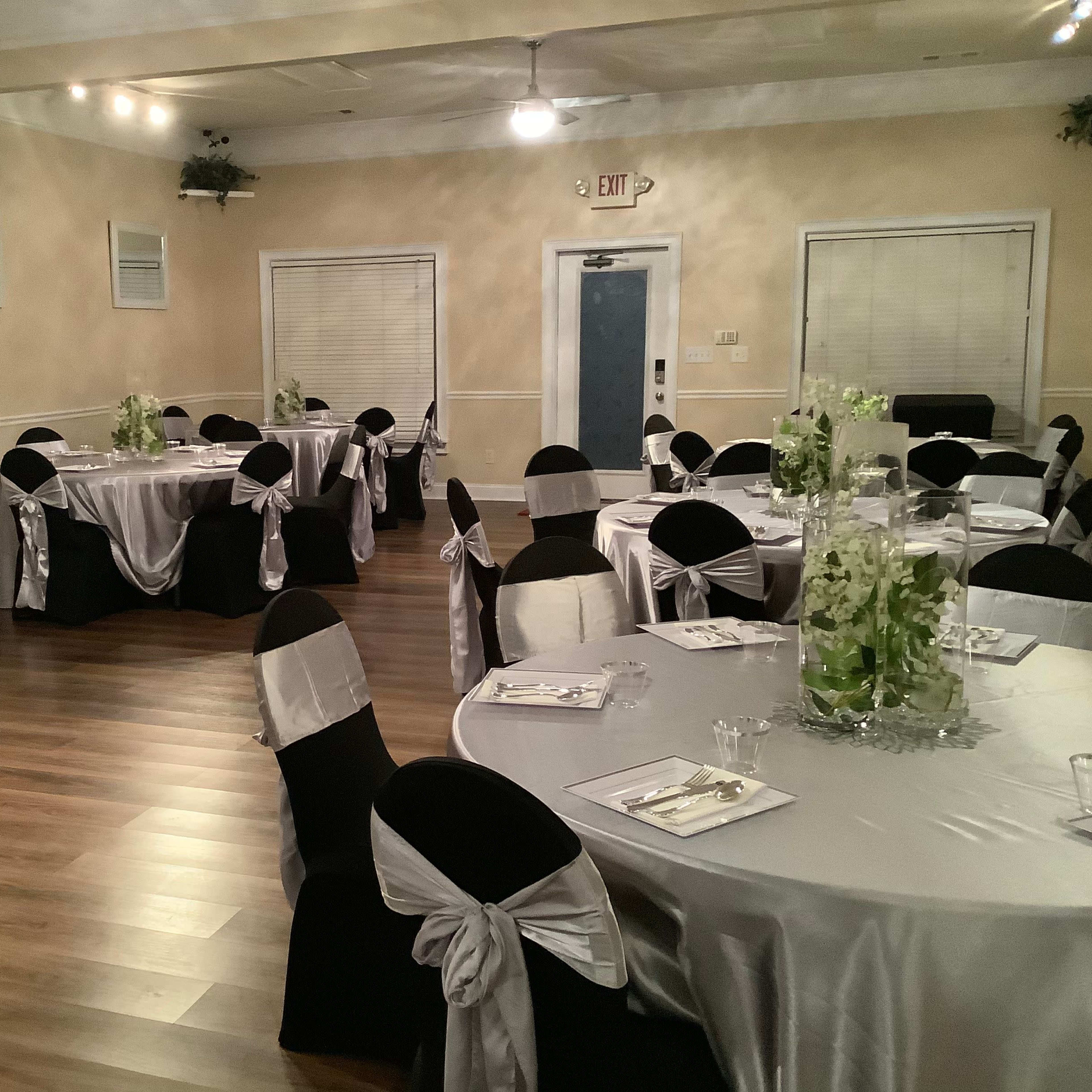 A decorated event space with round tables covered in silver and black linens, each set with white chairs adorned with silver bows.