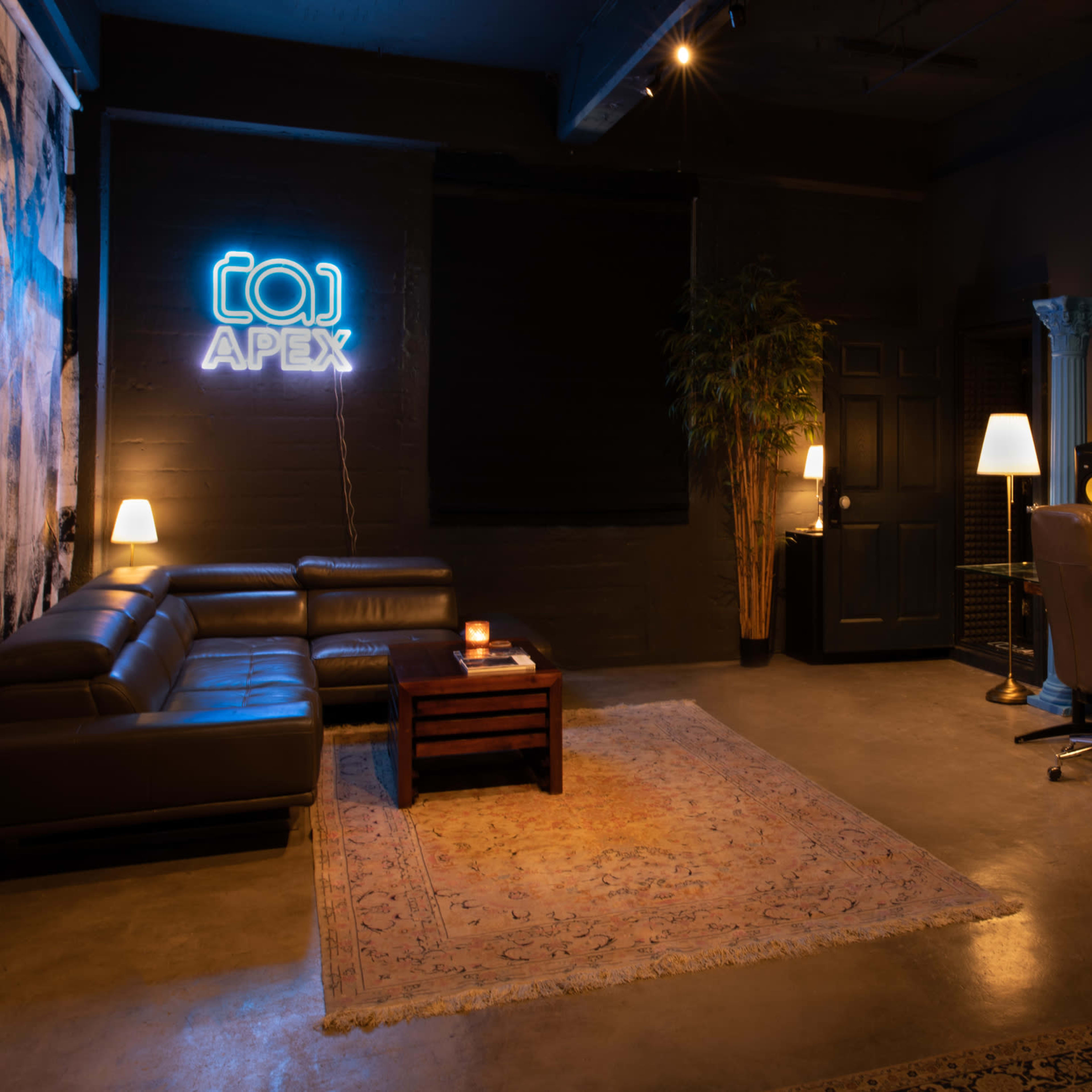 A dimly lit music studio features a leather sofa, a wooden table with a candle, a desk with recording equipment, and a neon "APEX" sign on the wall.
