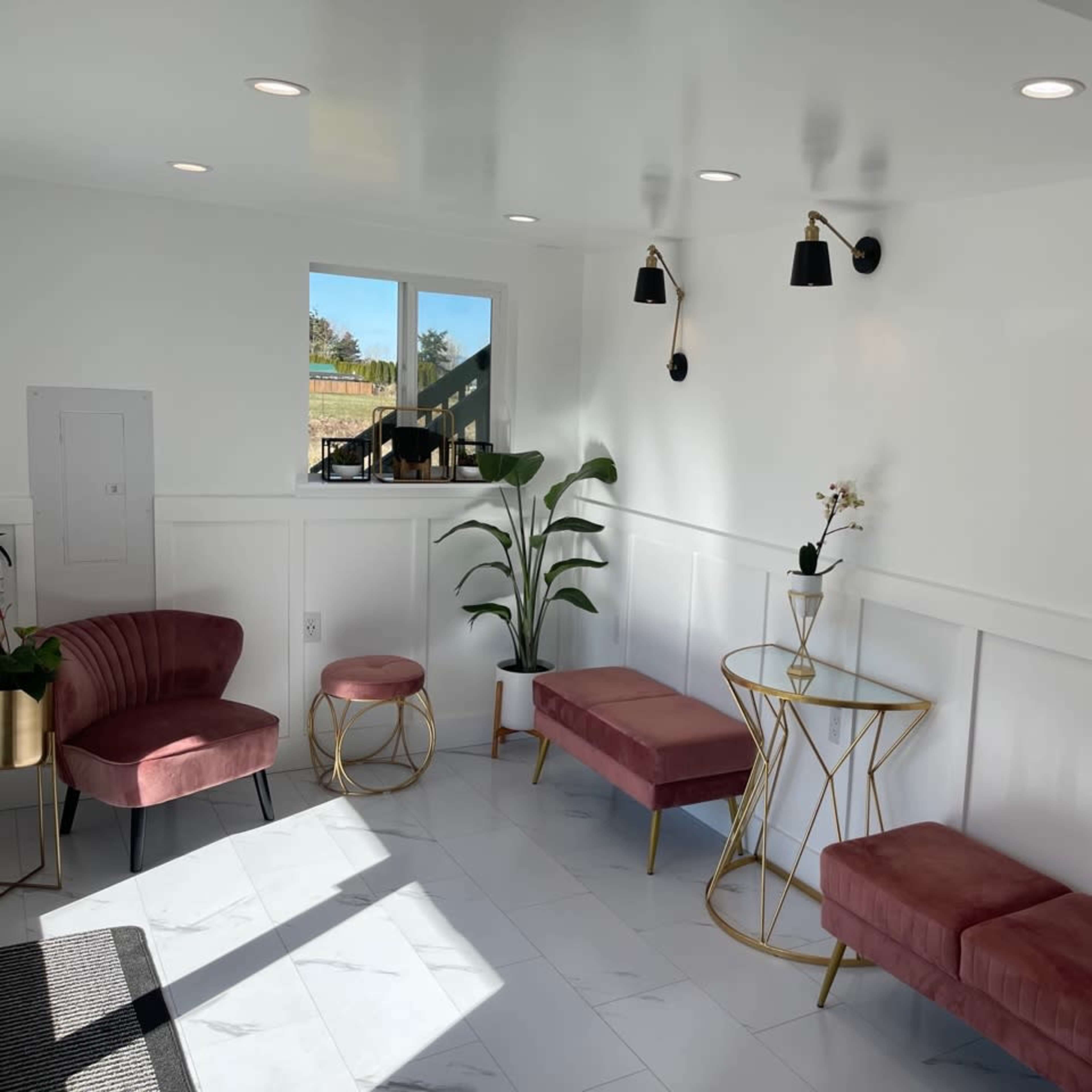 A bright room with white walls and a tiled floor, furnished with several pink seating options, a gold side table, and wall-mounted black lamps.