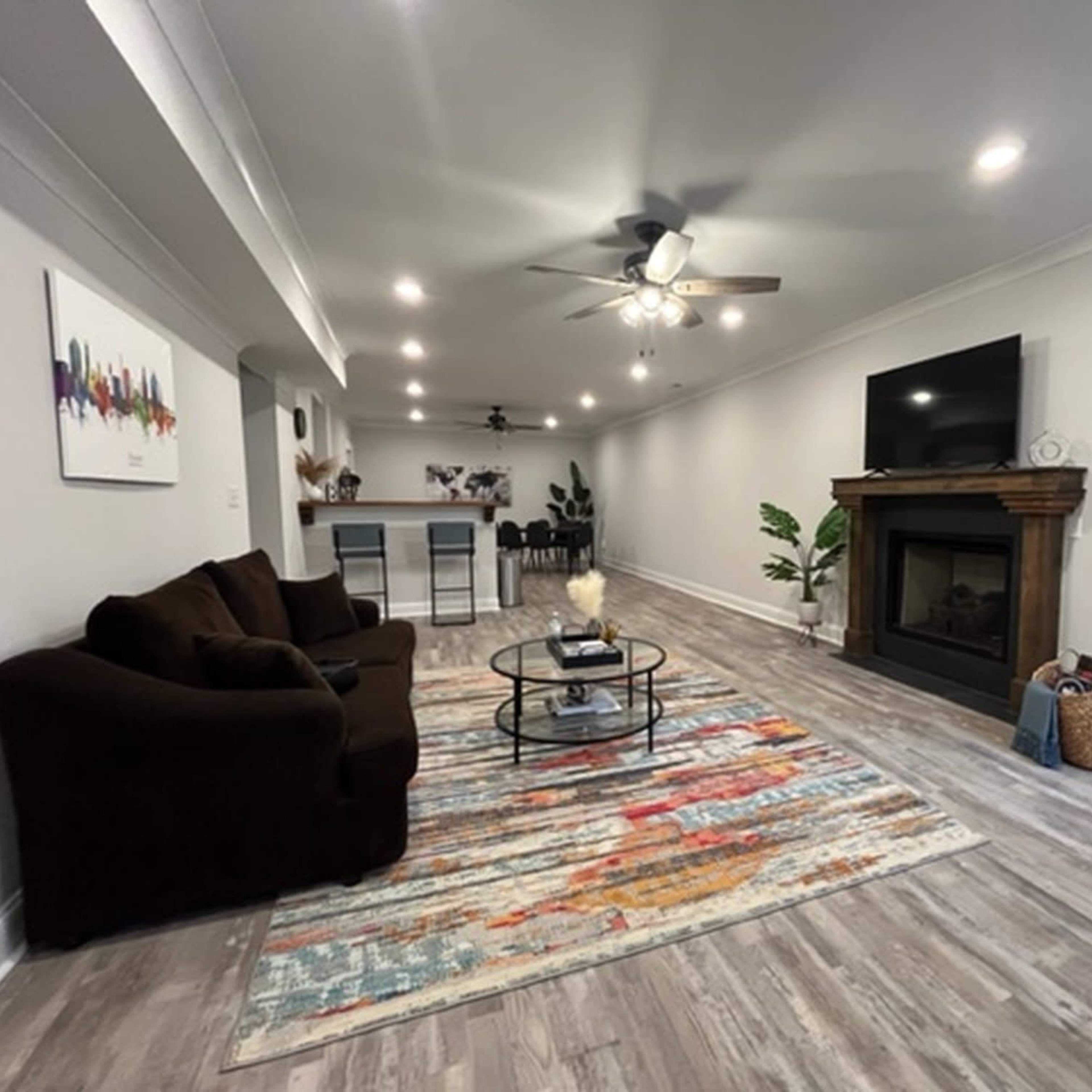 A modern living room with a brown couch, a circular coffee table on a colorful rug, and a wall-mounted TV above a fireplace, leading to a kitchen area in the background.