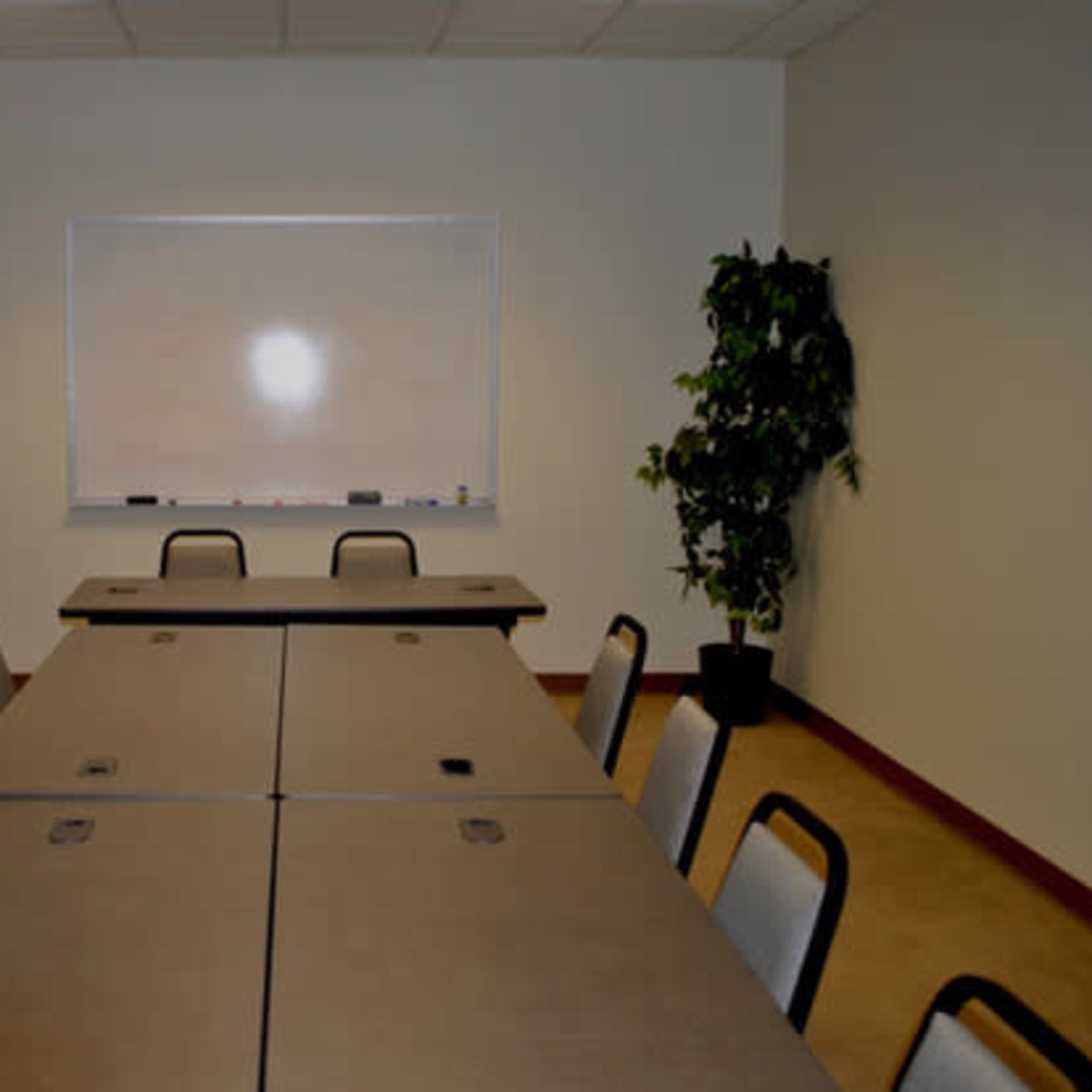 A conference room features several tables arranged in a rectangular formation, with chairs, a whiteboard, and a potted plant in the corner.