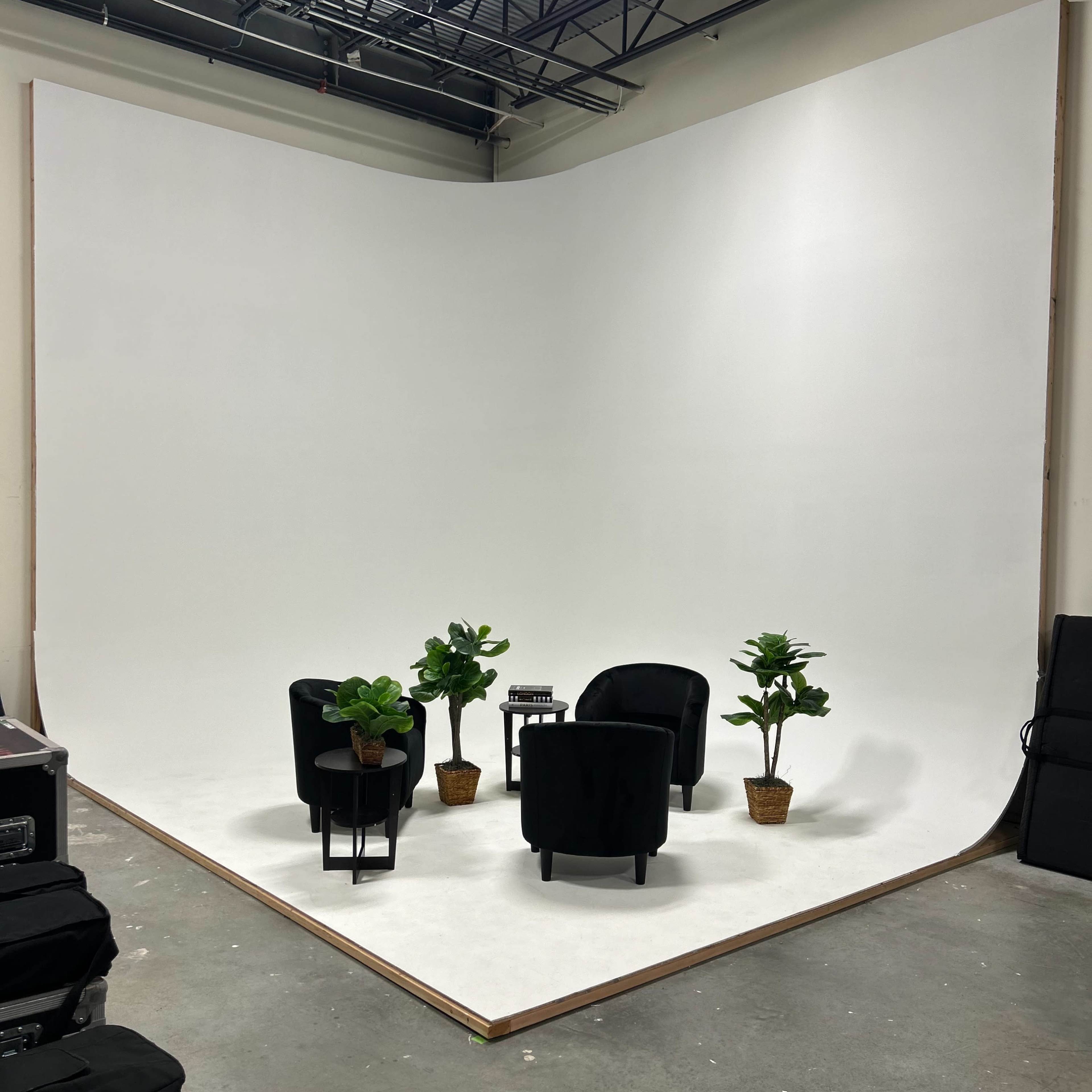 A simple studio setup with two black chairs, a small table, and potted plants against a large, white backdrop.