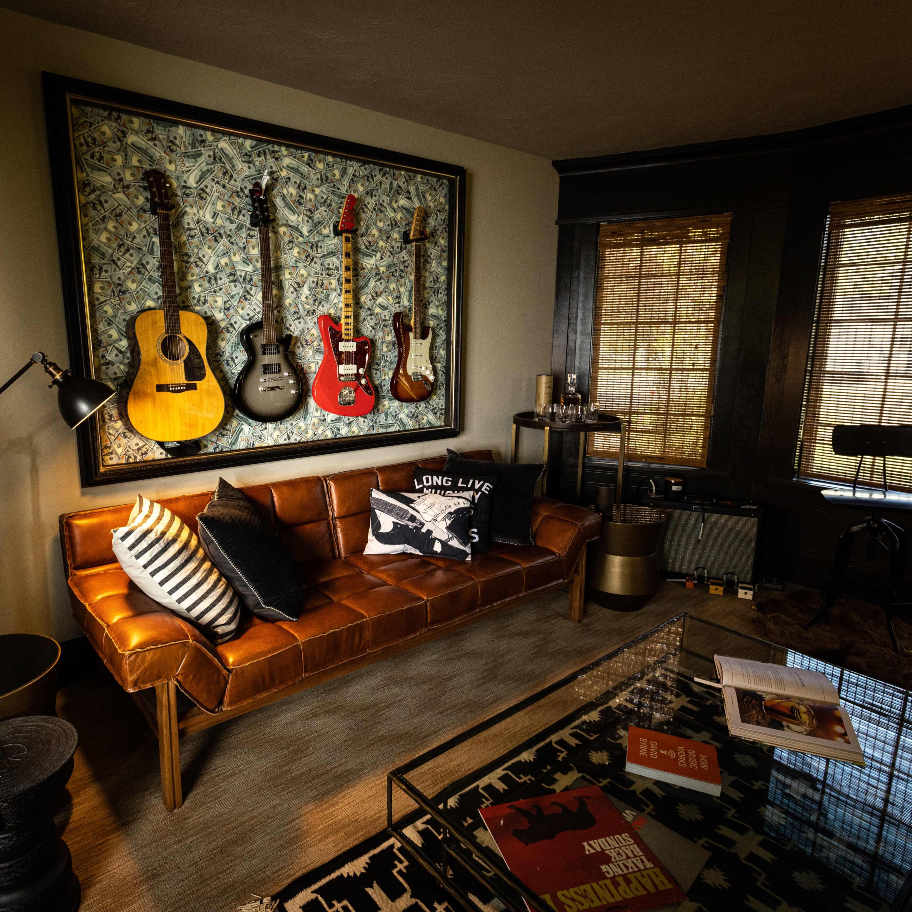 A living room featuring a brown leather couch, a wall adorned with four guitars, and a coffee table with books and decor.