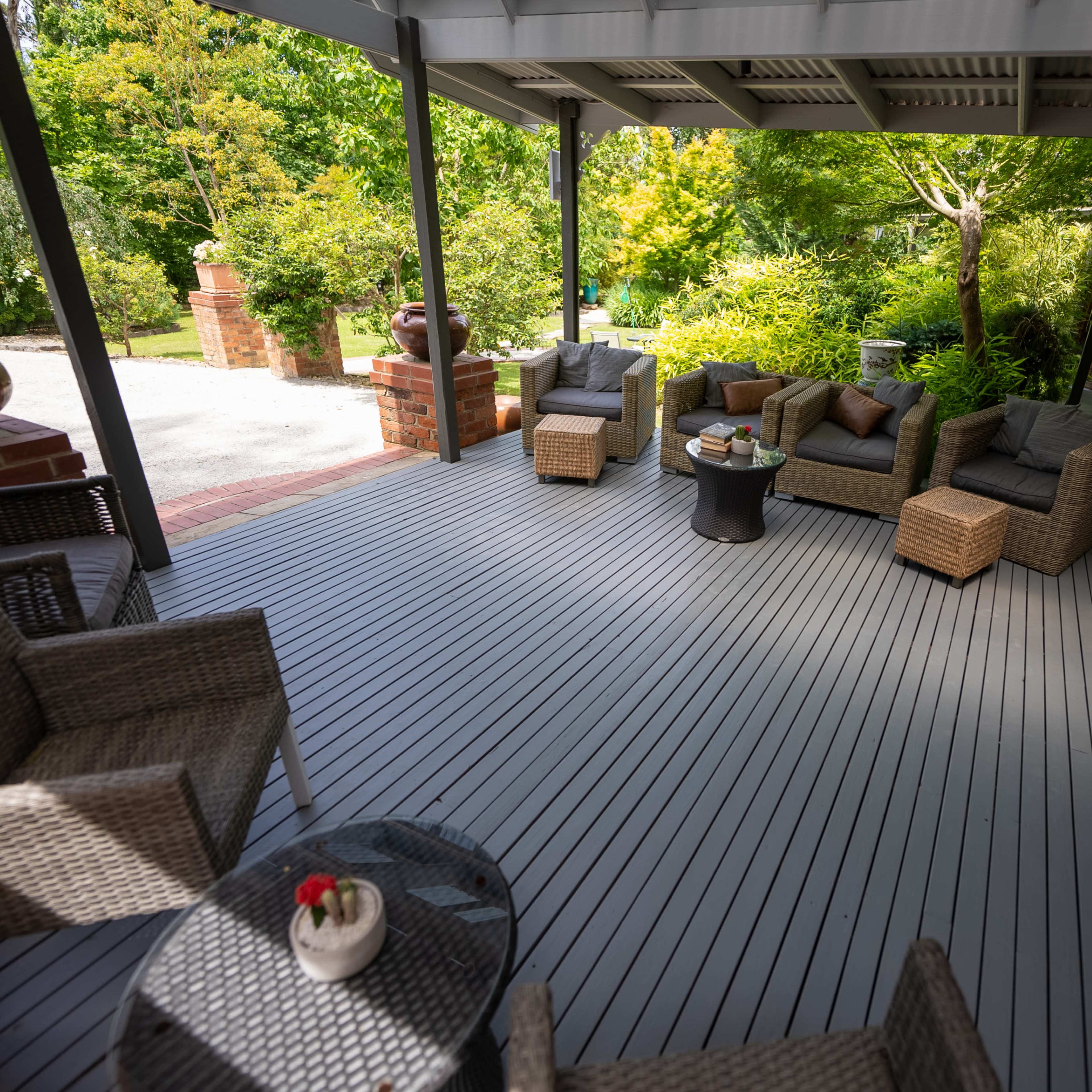 A spacious porch features a seating arrangement with wicker chairs, a small table, and green foliage in the background.