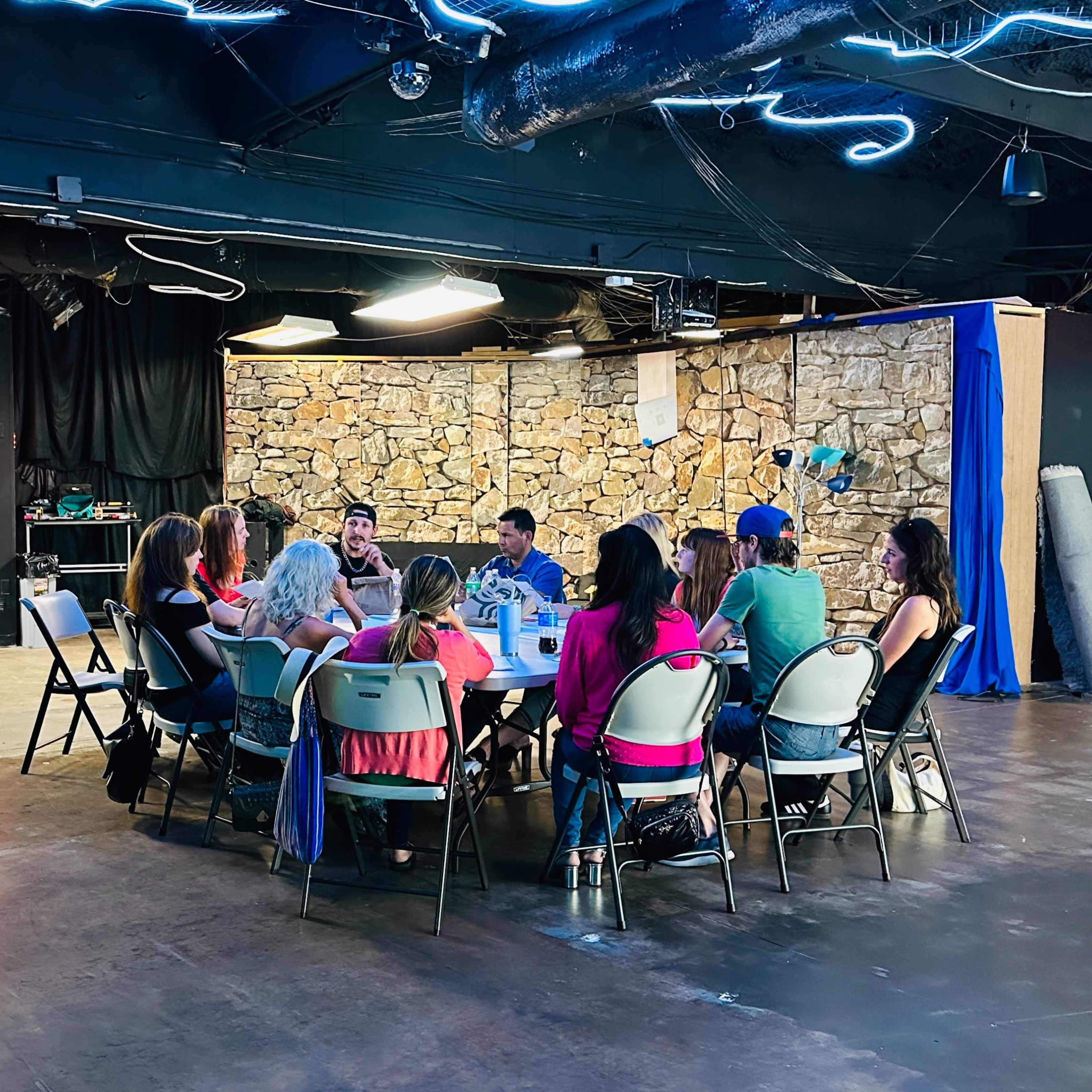 A group of people sits around a circular table in a spacious, unfinished room with stone walls and bright overhead lights.