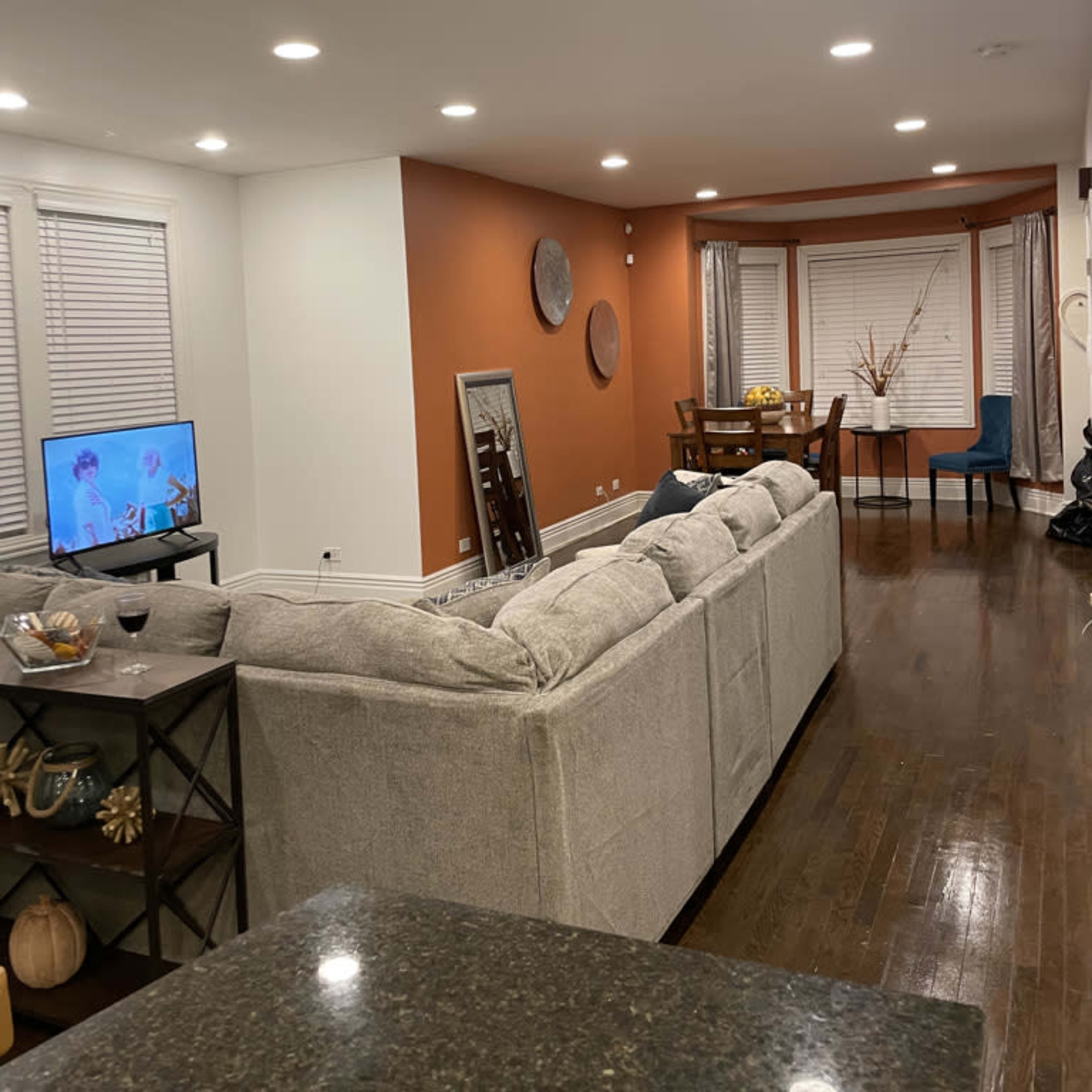 A living room features a gray sofa facing a television, with an adjacent dining area and warm-colored accent wall.