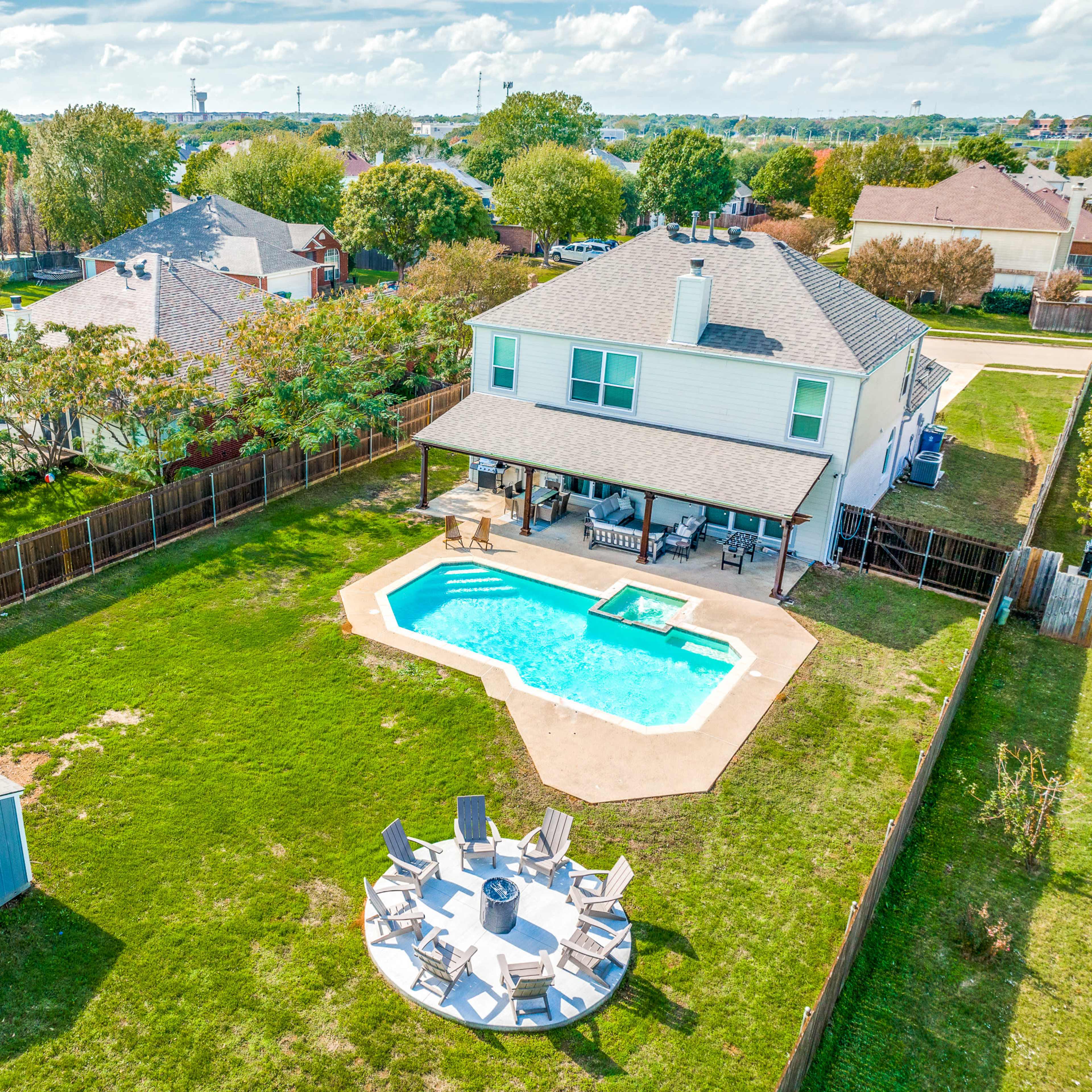 A backyard with a swimming pool, seating area, and a small shed, surrounded by a wooden fence and residential buildings in the background.