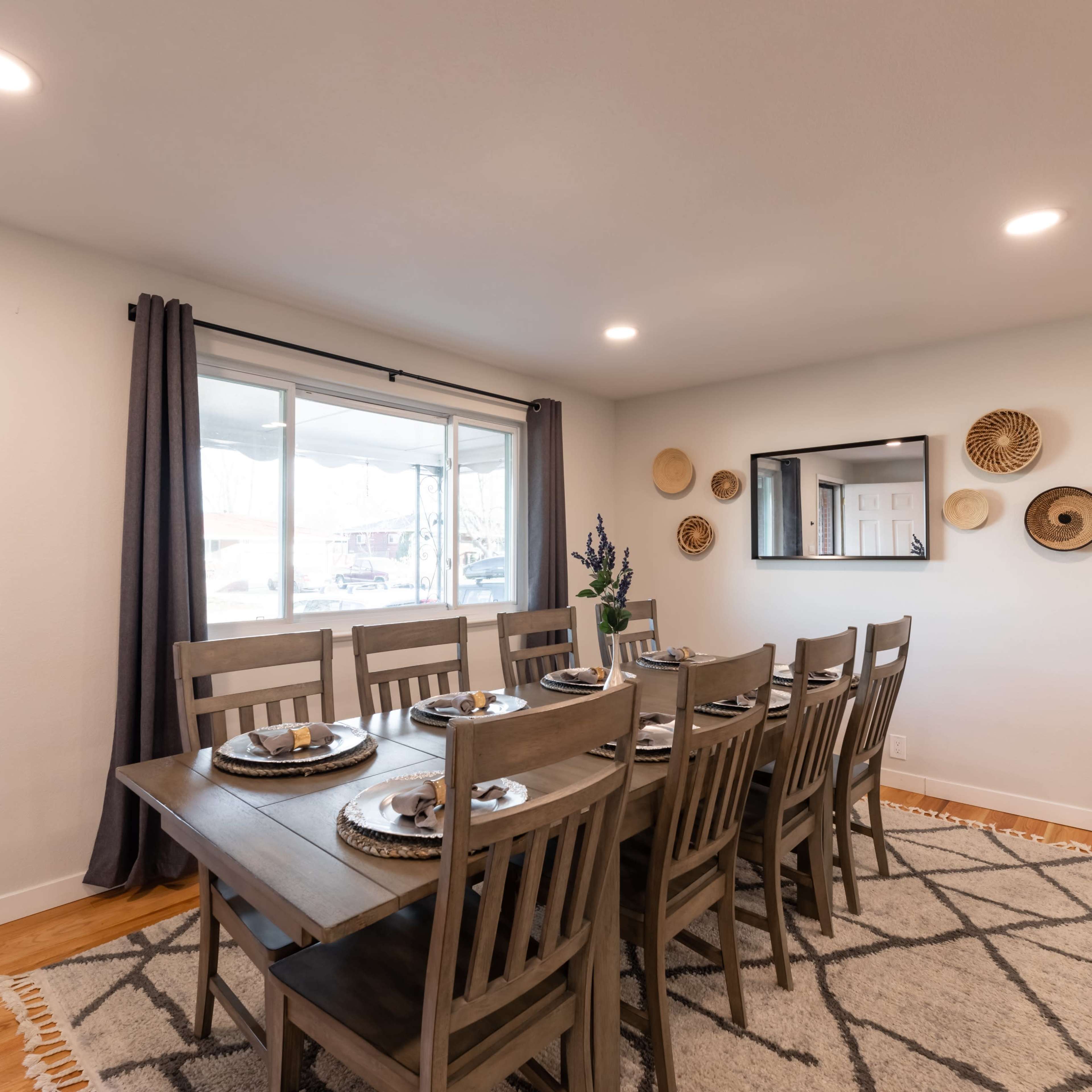 A spacious dining area features a long wooden table set for eight, with natural light coming through a large window and decorative wall hangings.