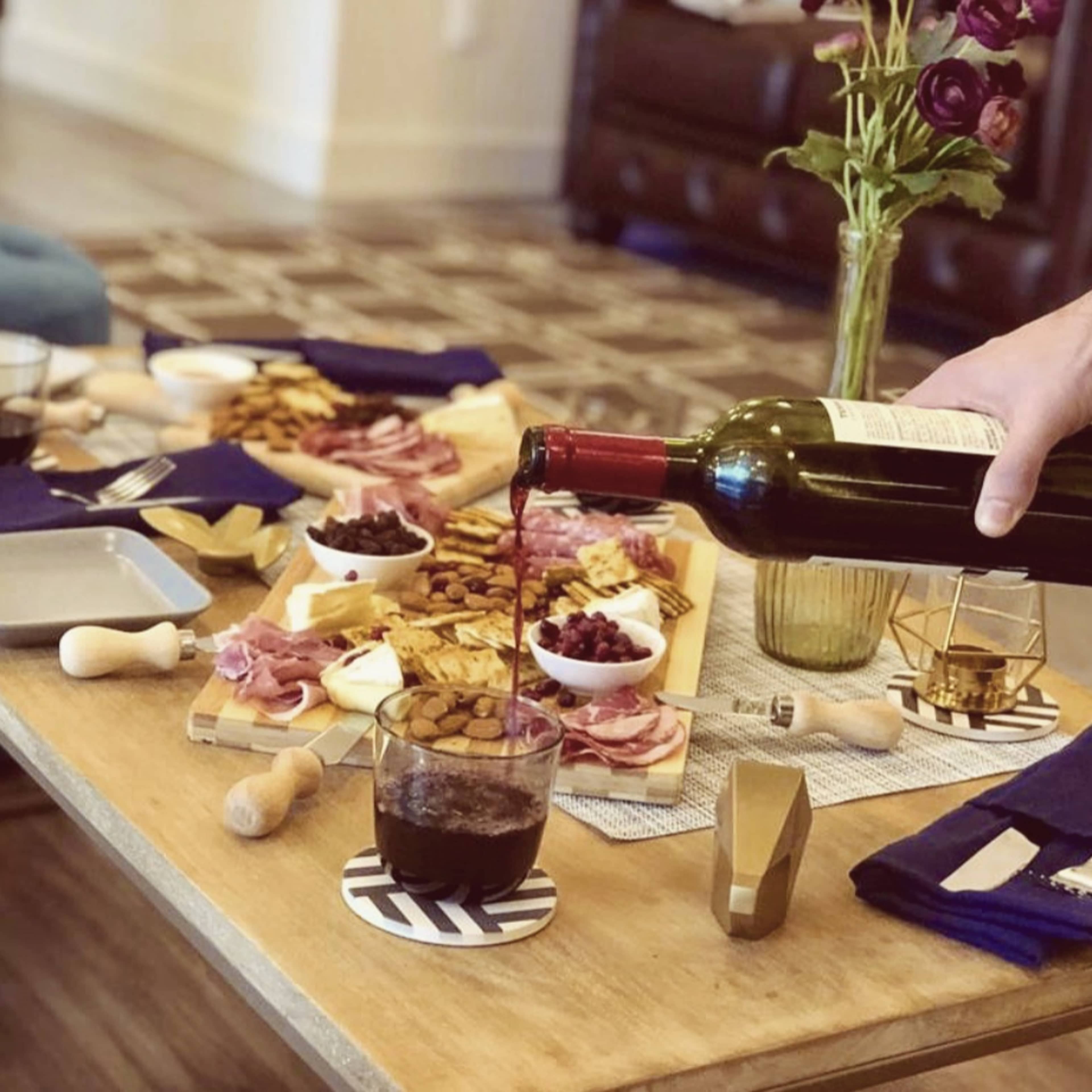 A hand pours red wine from a bottle into a glass on a wooden table set with a variety of meats, cheeses, fruits, and decorative accessories.