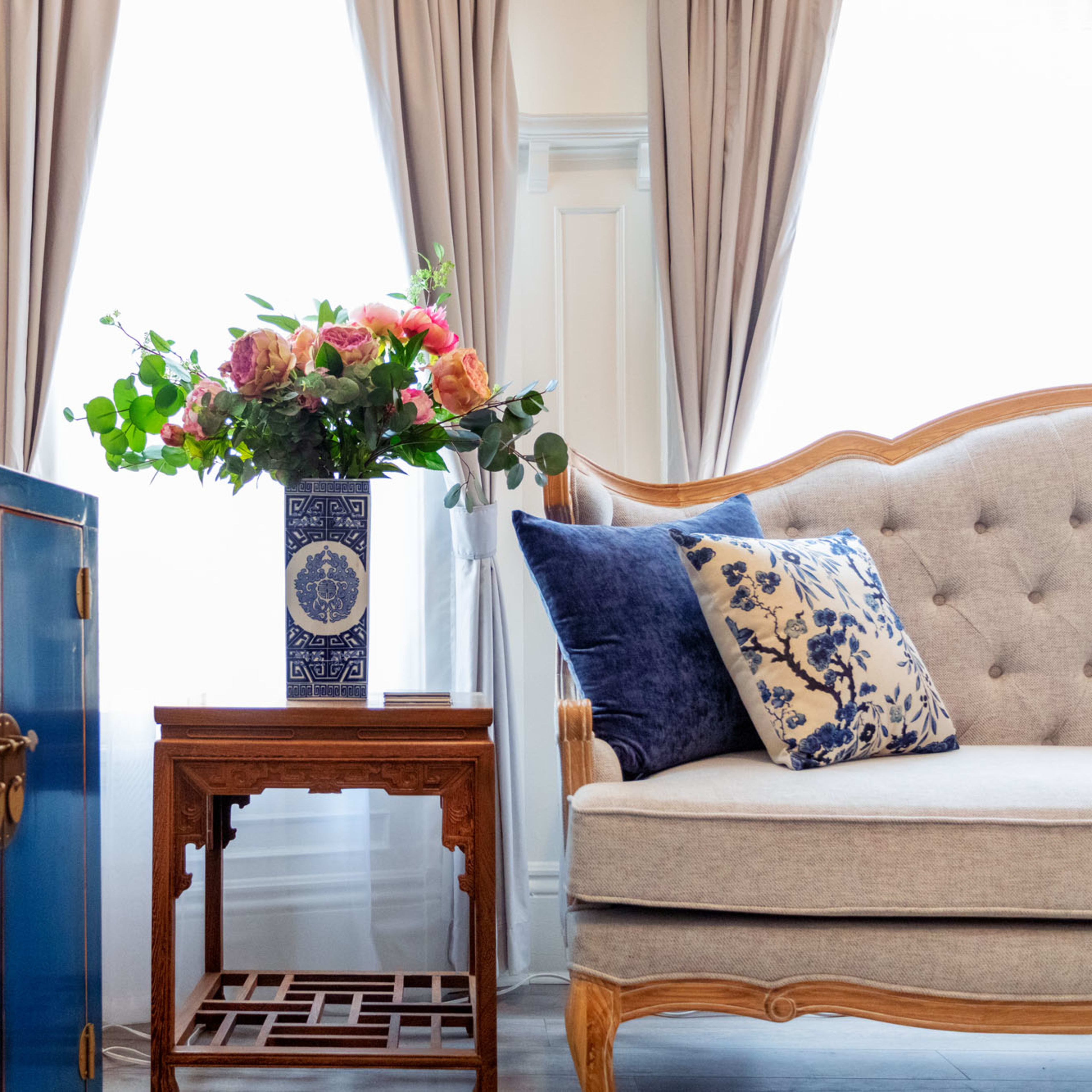 A light-filled room features a blue cabinet, an ornate wooden side table, a gray tufted sofa with decorative pillows, and a vase of flowers on the table.