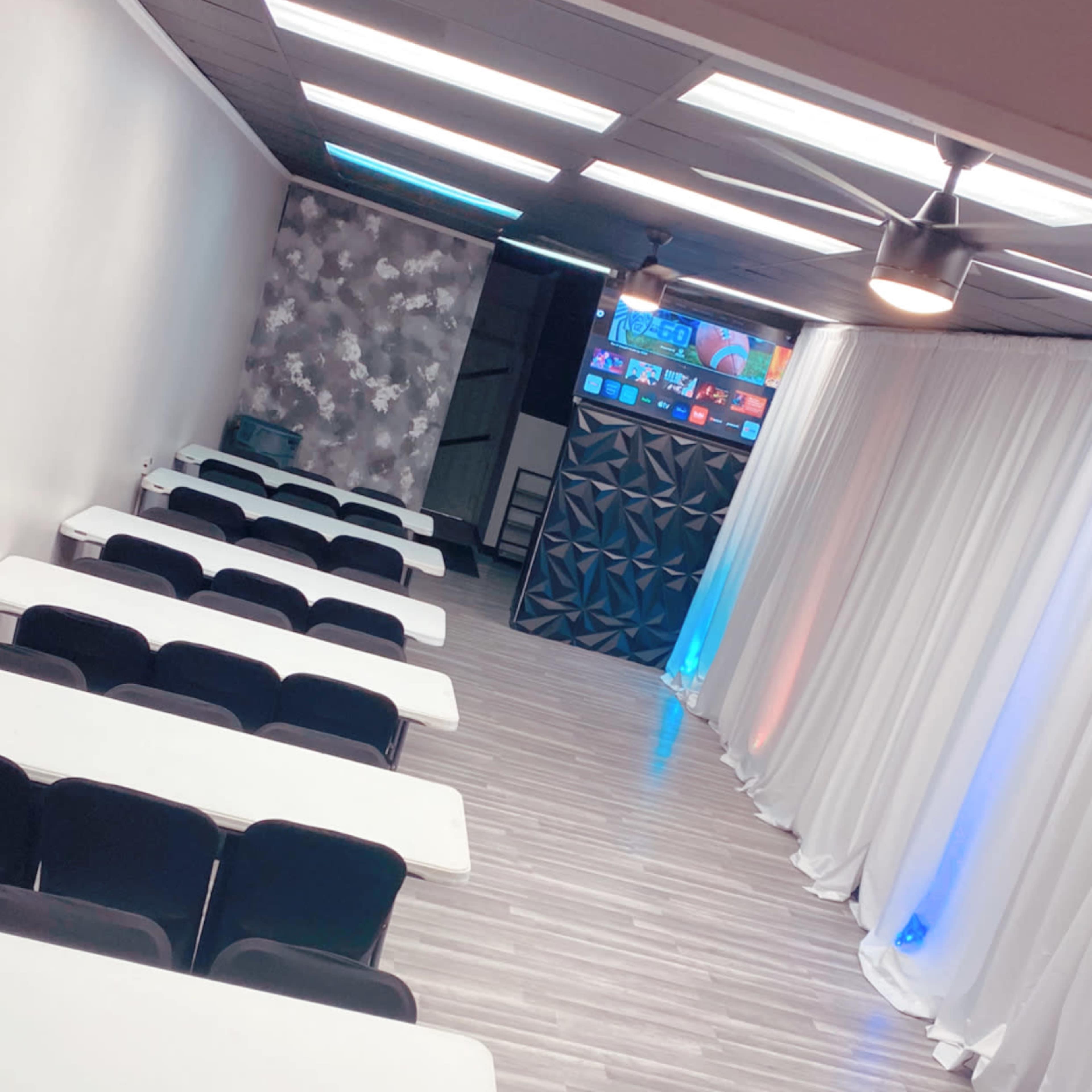 A spacious room arranged with several rows of tables and chairs, accompanied by a large screen at the far end and illuminated curtains along the walls.