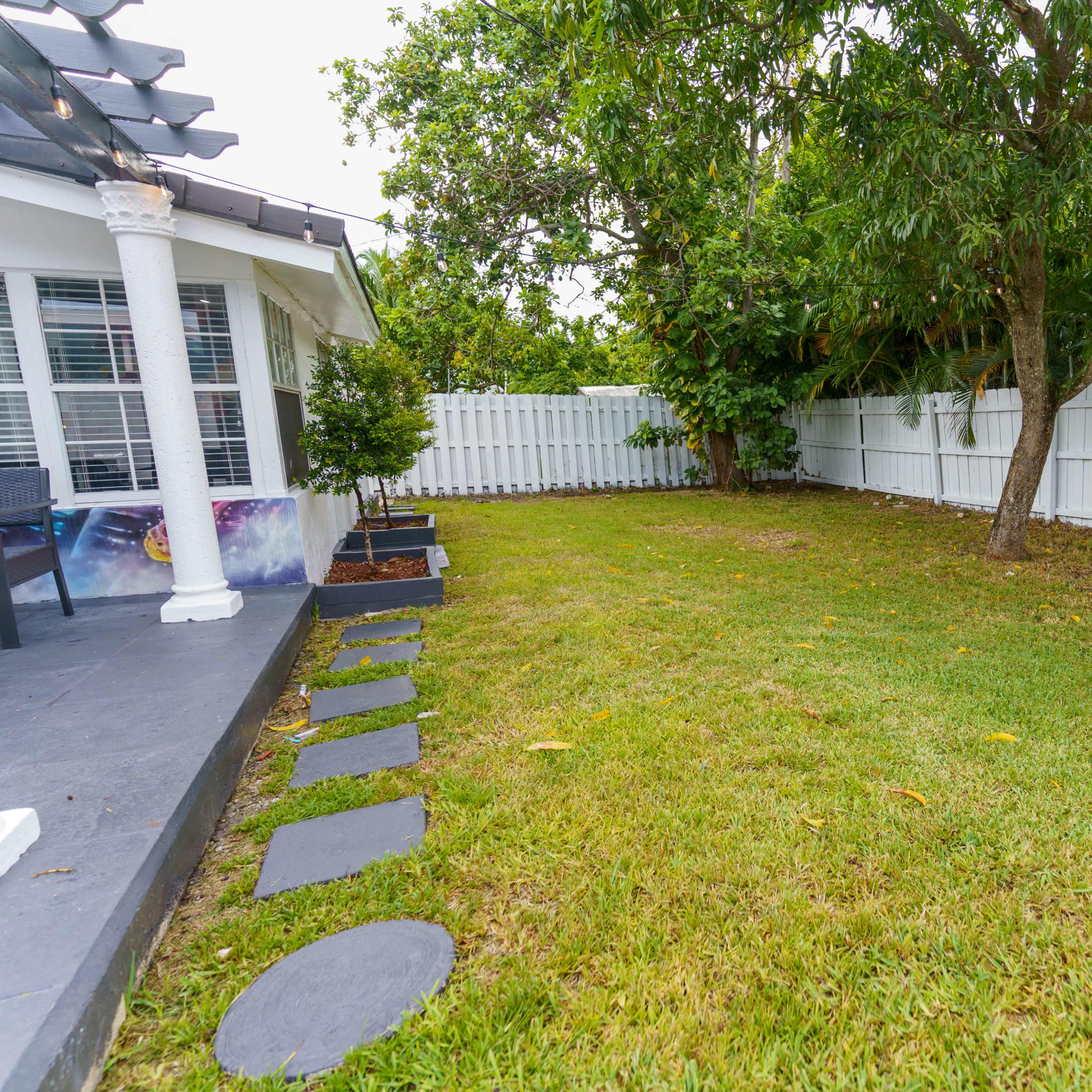 A grassy backyard with a white wooden fence, a stone walkway, and a small tree.