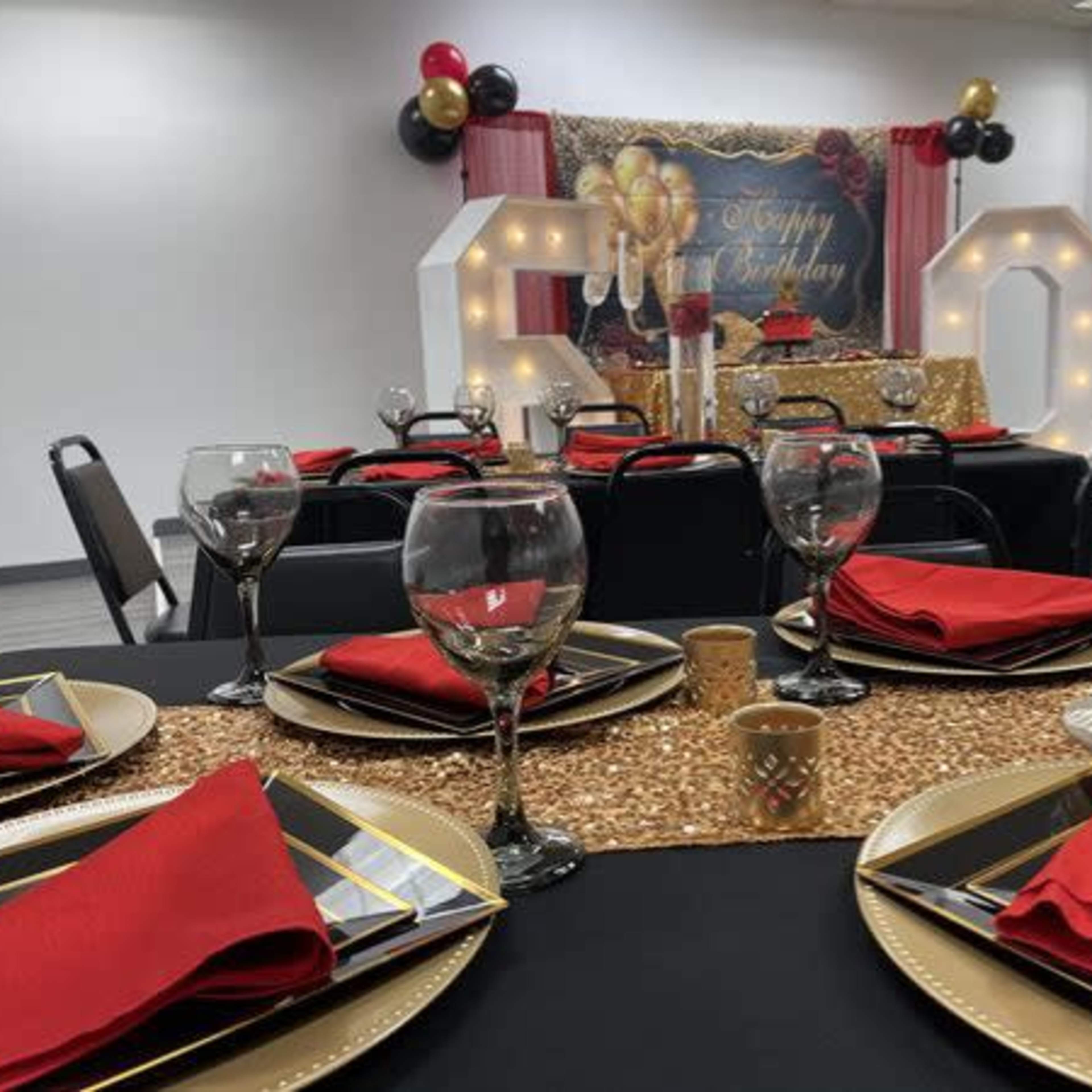 A decorated event space with tables set in black and gold featuring red napkins, glassware, and a backdrop adorned with balloons and a festive sign.