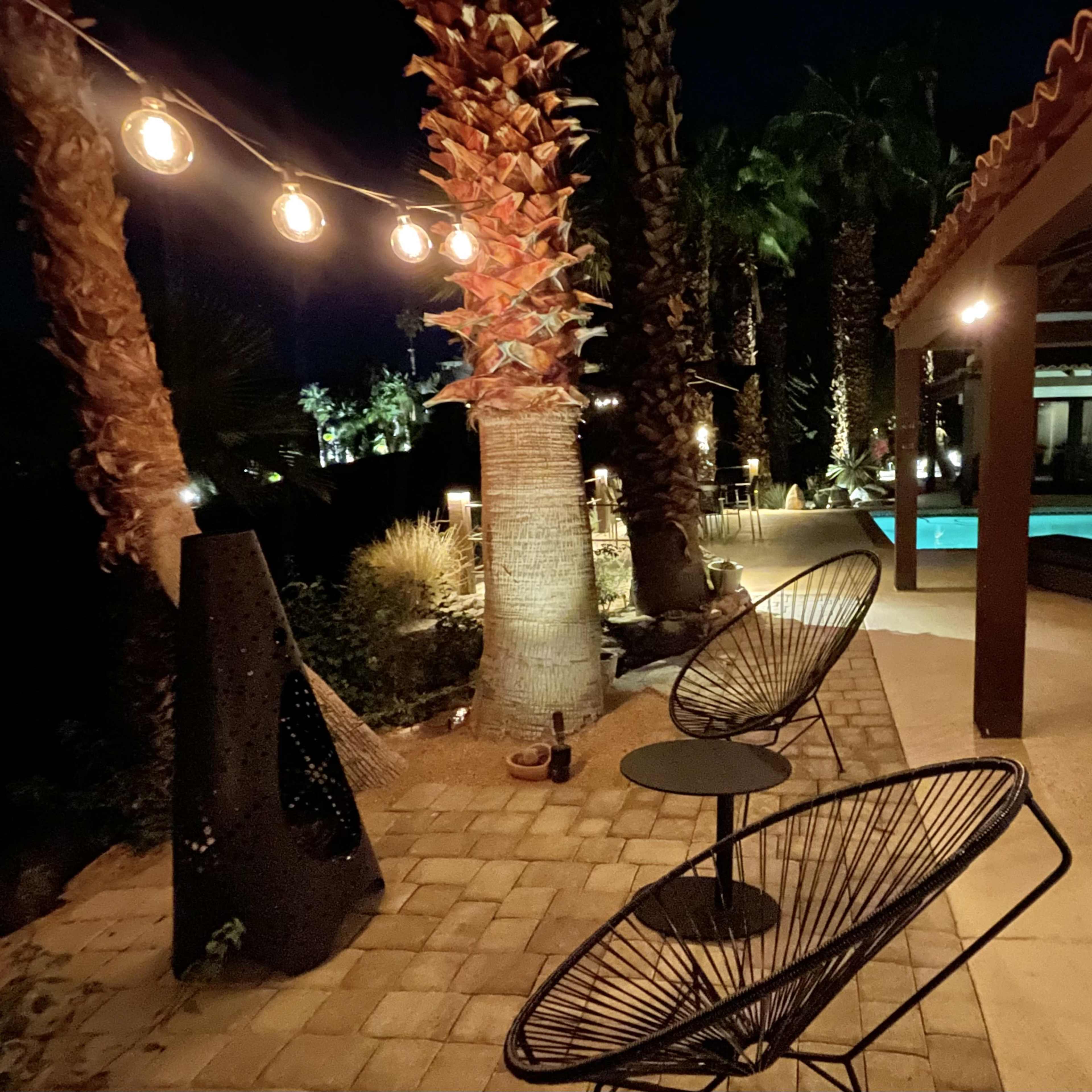 A patio area at night with two chairs, a small table, palm trees, and a swimming pool visible in the background, illuminated by string lights.