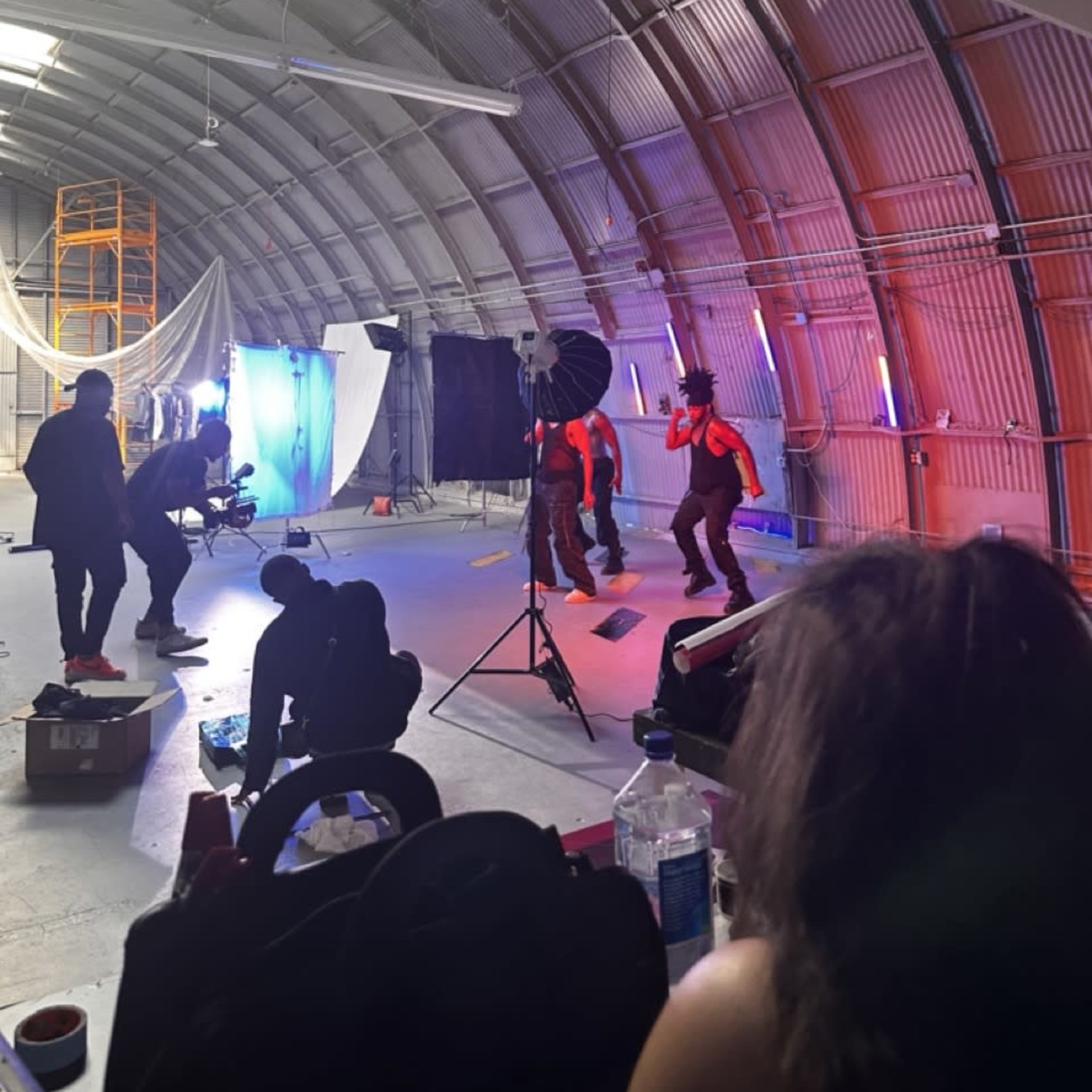A group of people in a studio setting are filming a scene with colorful lighting and various camera equipment.