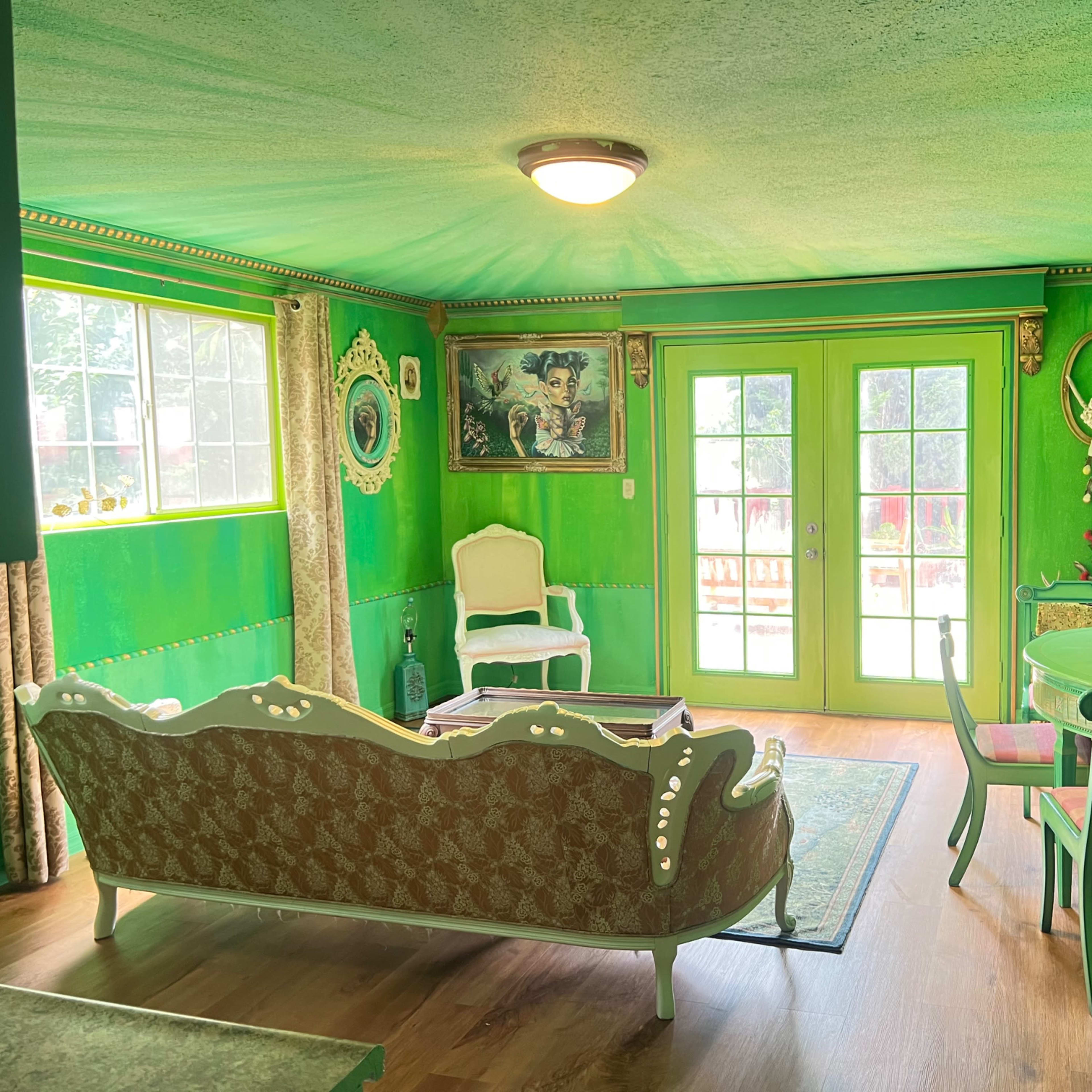 The room features green walls and ceiling, a patterned couch, and a small dining table near glass doors leading to an outdoor area.