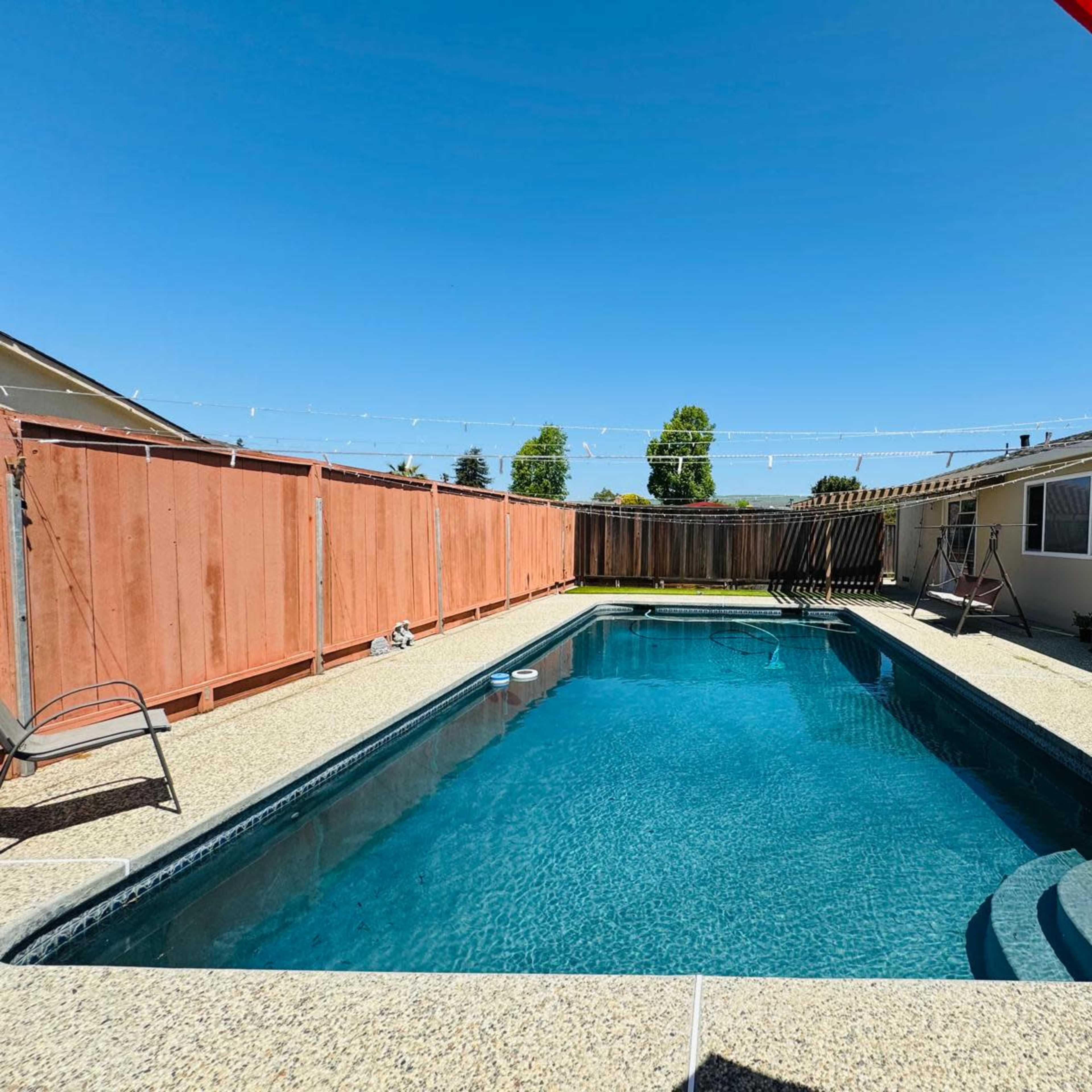 A clear blue swimming pool is situated in a fenced backyard, surrounded by a concrete deck and featuring a patio umbrella.