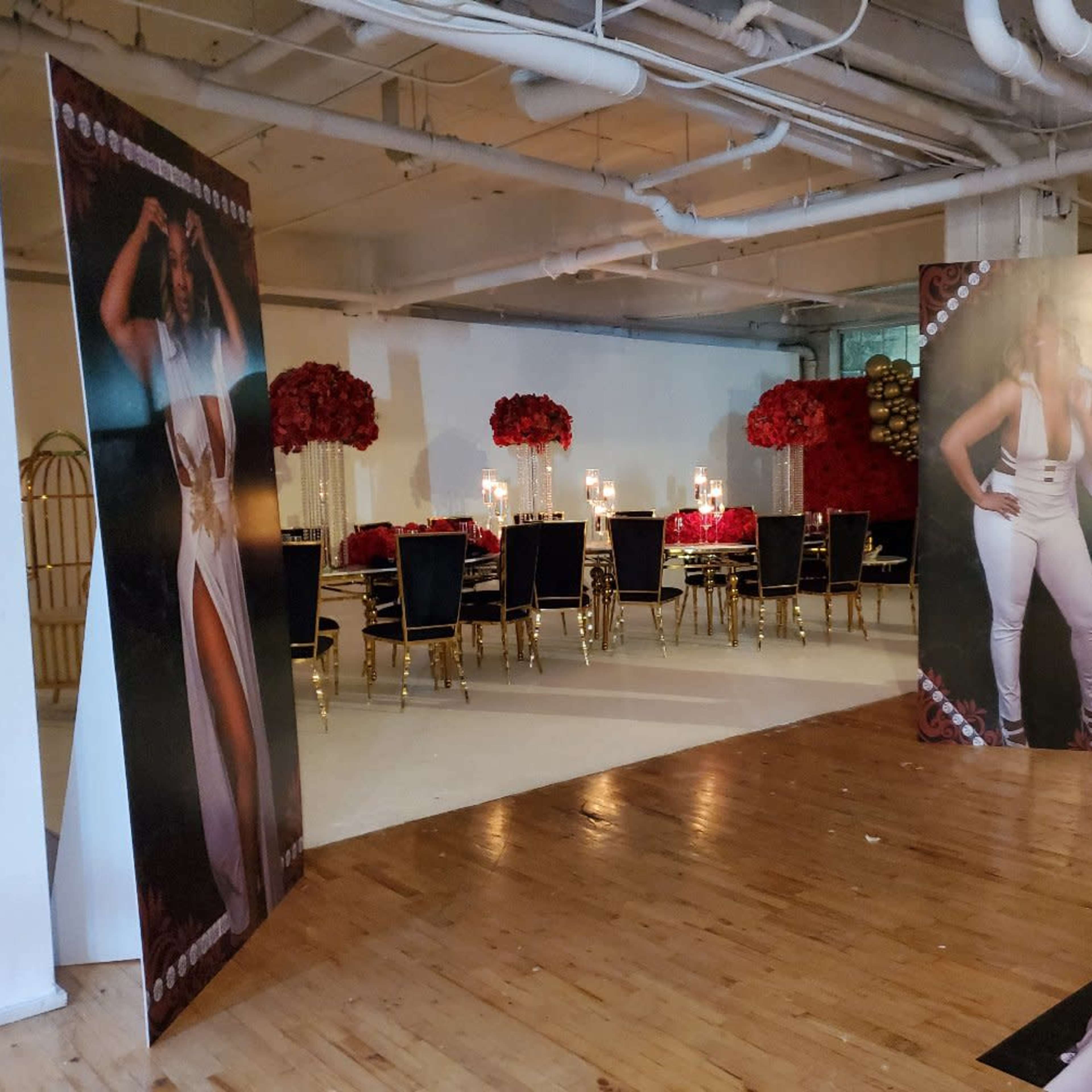 A decorated event space featuring tall floral arrangements, a long dining table with candles, and large banners displaying images of a woman.