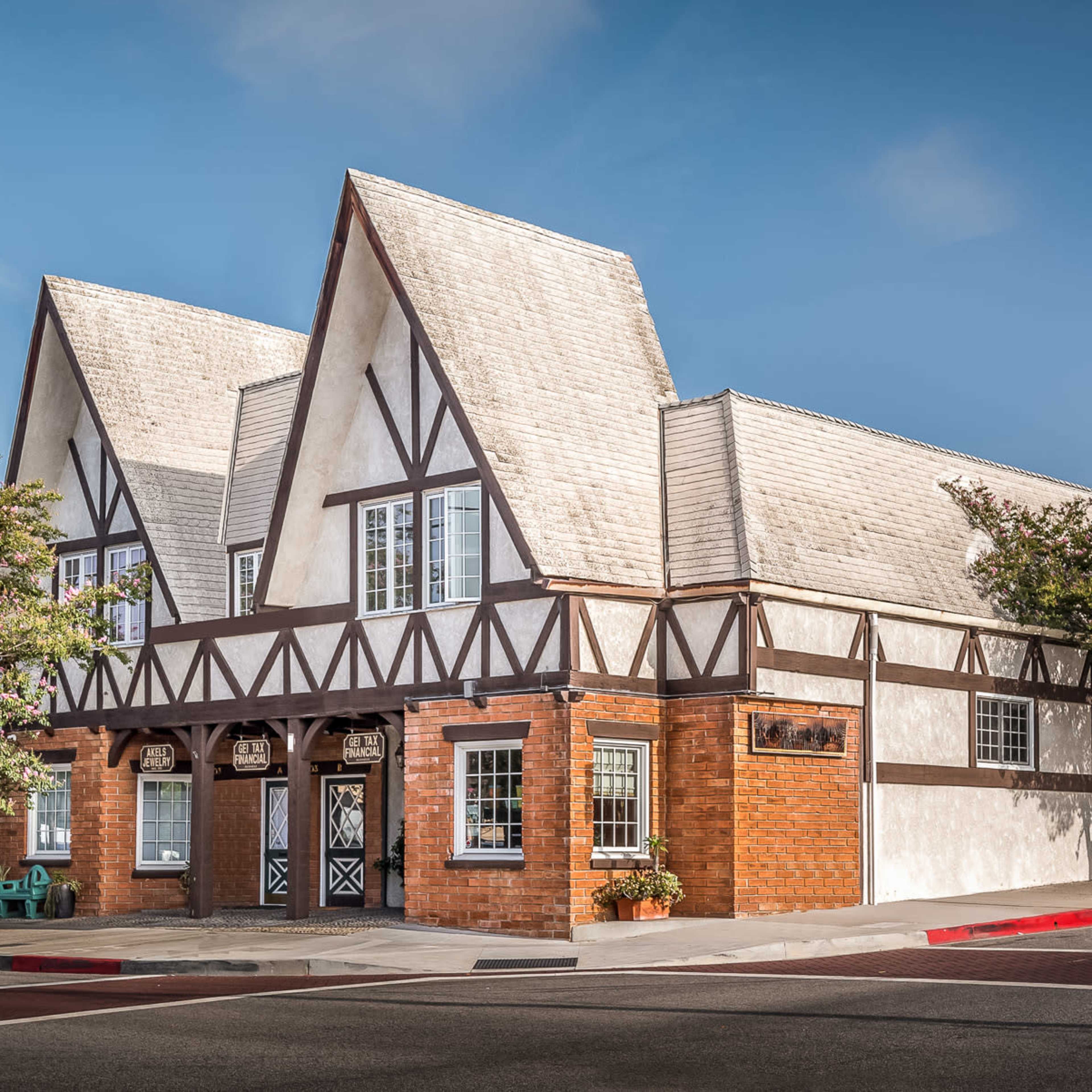 A two-story building with a gabled roof and timber framing is situated on a street corner, surrounded by trees and planters.