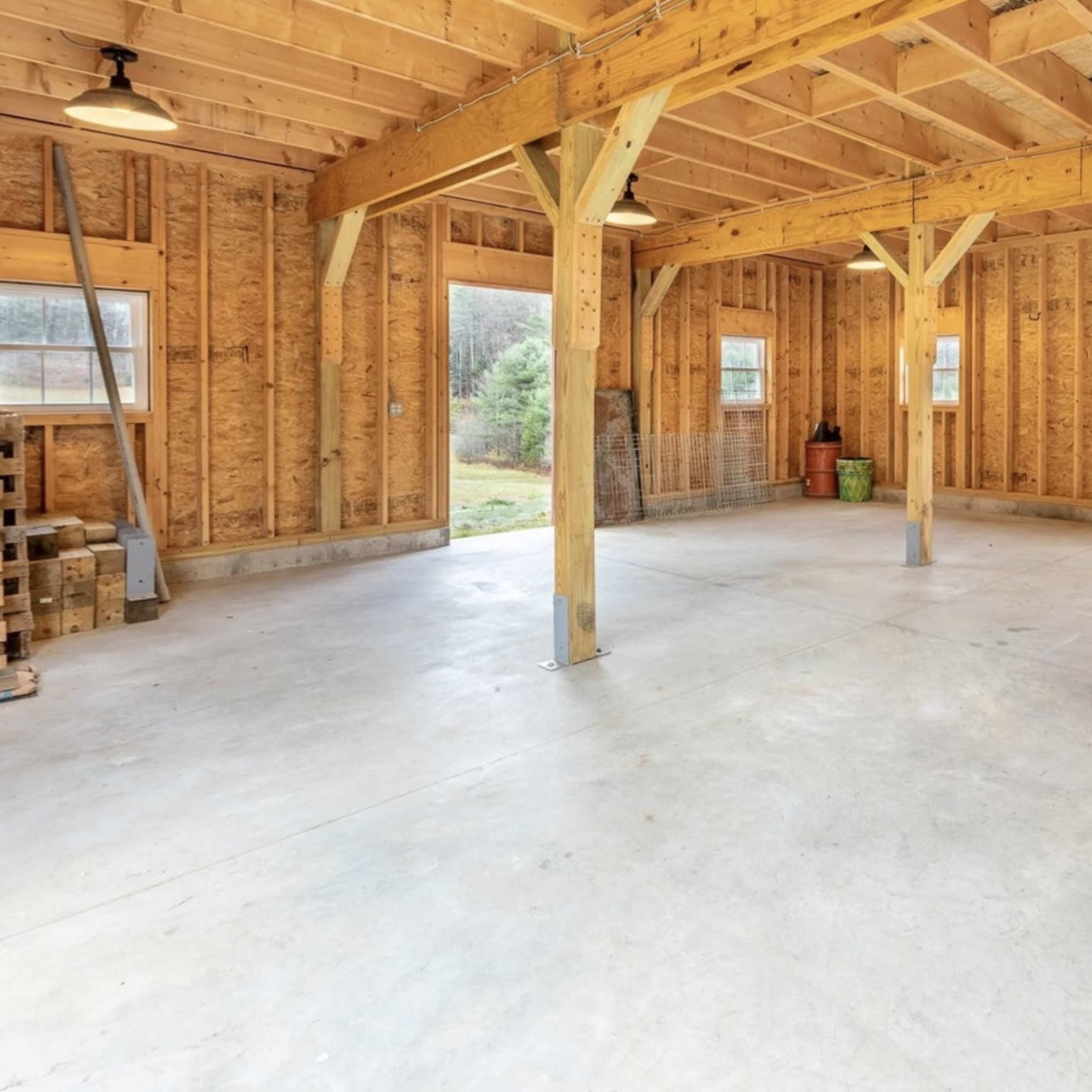 An empty wooden structure with exposed beams and a concrete floor, illuminated by overhead lights.