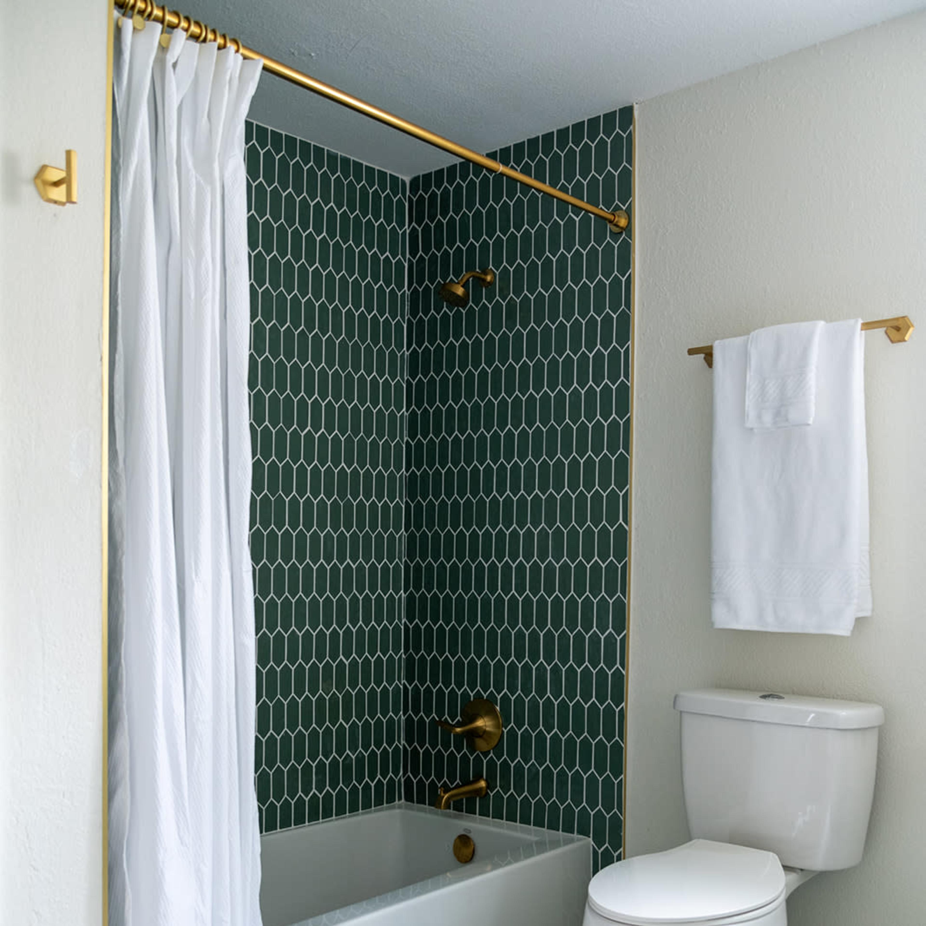 A bathroom with a bathtub surrounded by green honeycomb tile, a shower curtain, a gold shower fixture, and a white toilet.