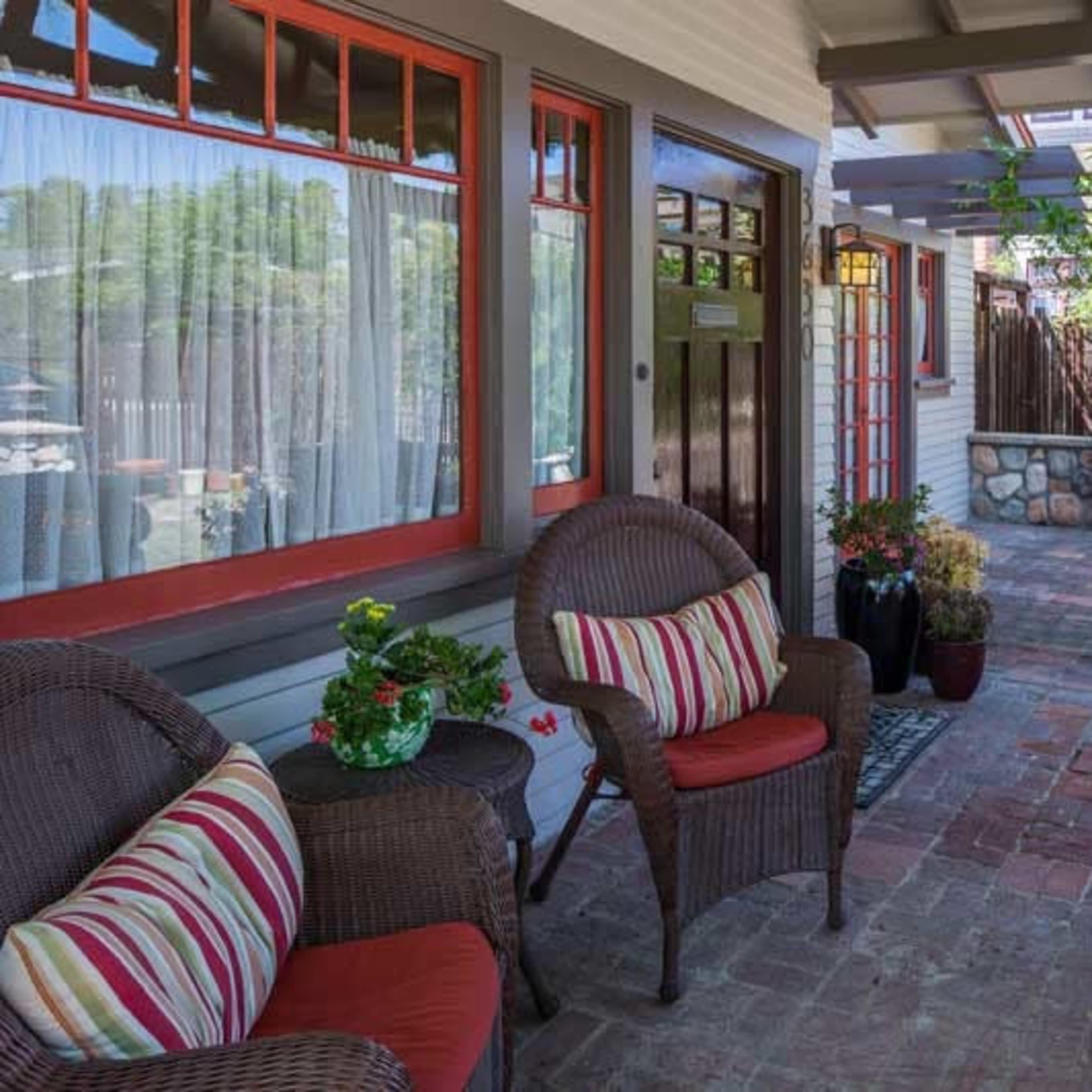A porch with two wicker chairs adorned with striped cushions, brick flooring, and potted plants against a backdrop of a wooden door and a garden.