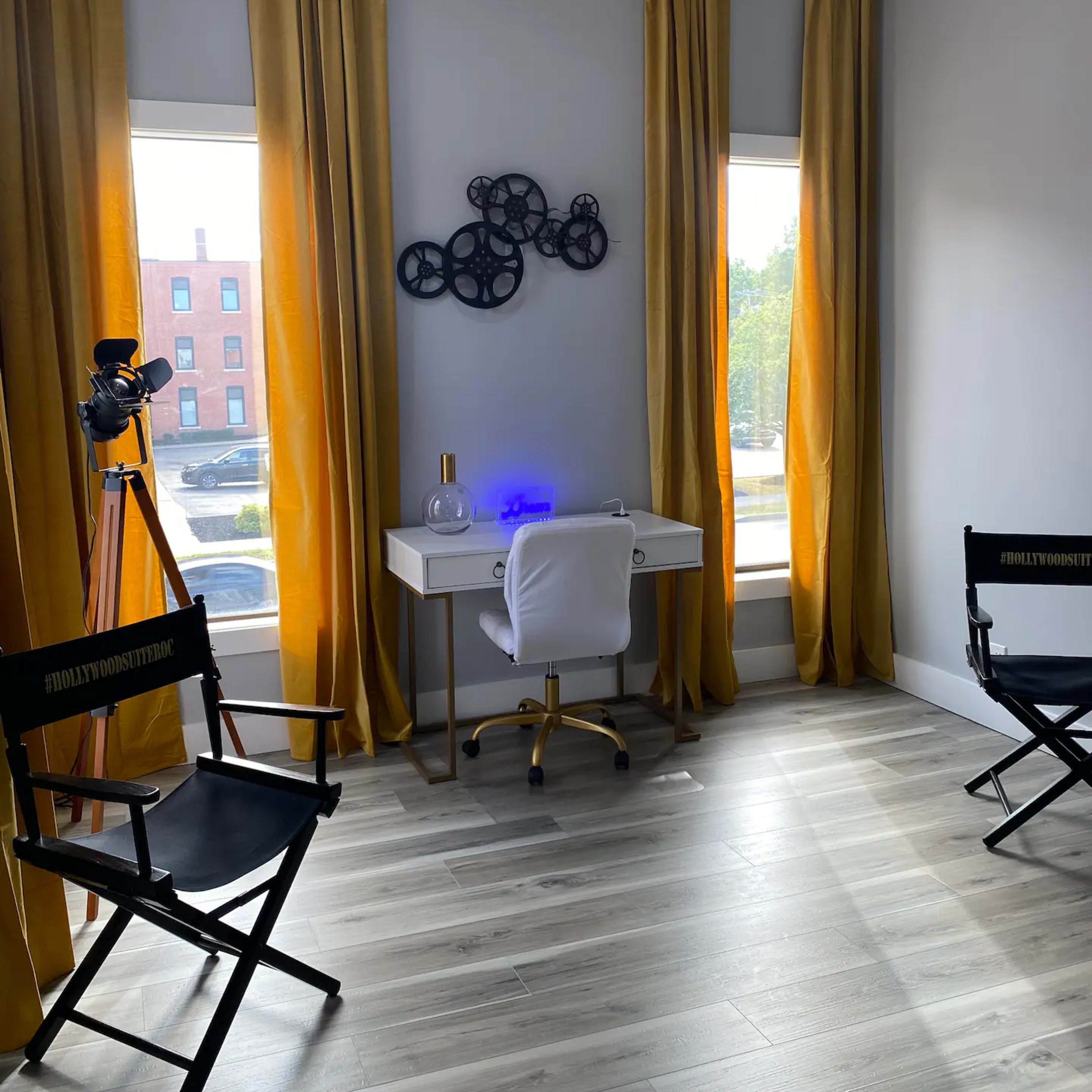 A well-lit room features a white desk and chair, two director's chairs, large windows with yellow curtains, and decorative wall art.