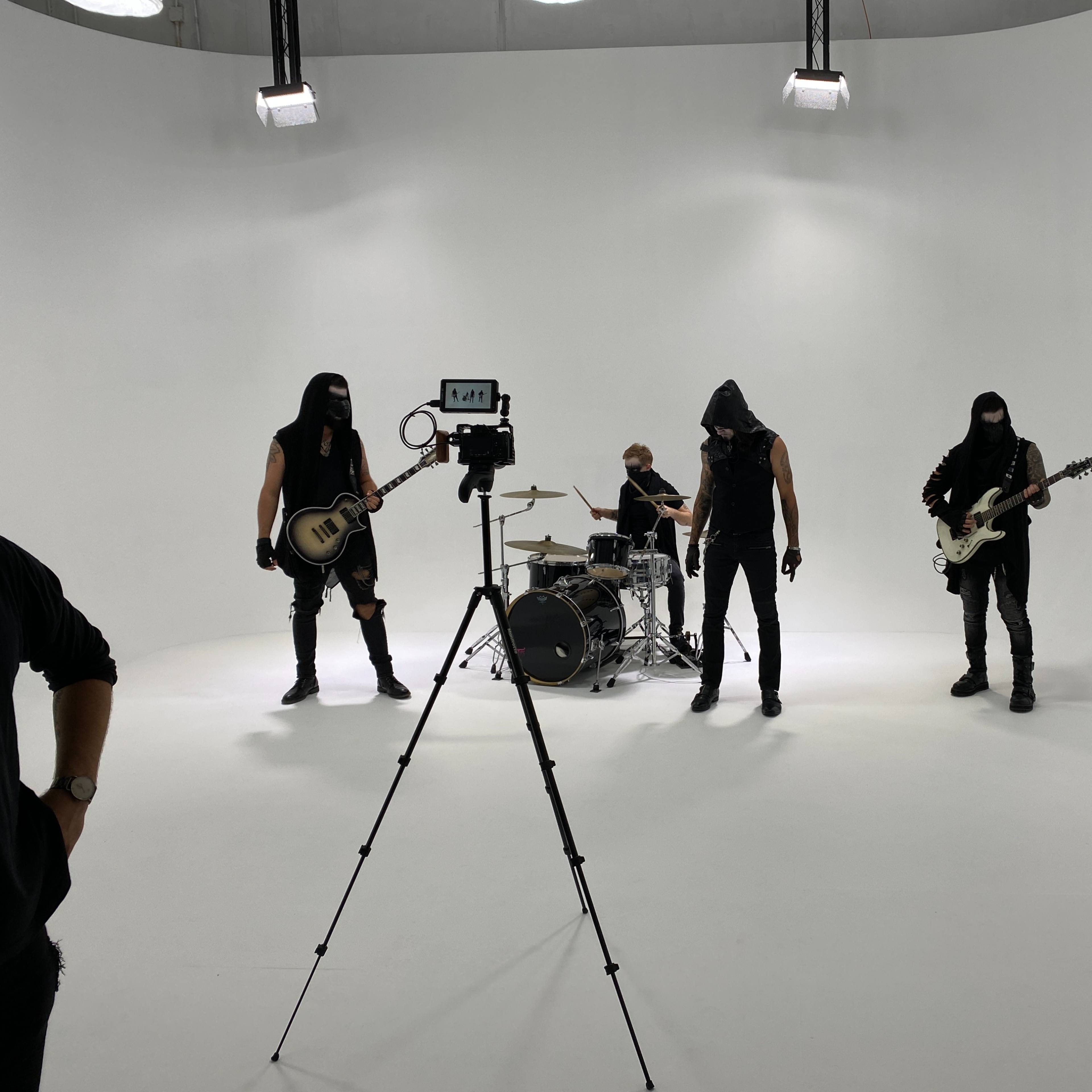 Four musicians dressed in dark clothing perform on a white stage, with one person filming them.