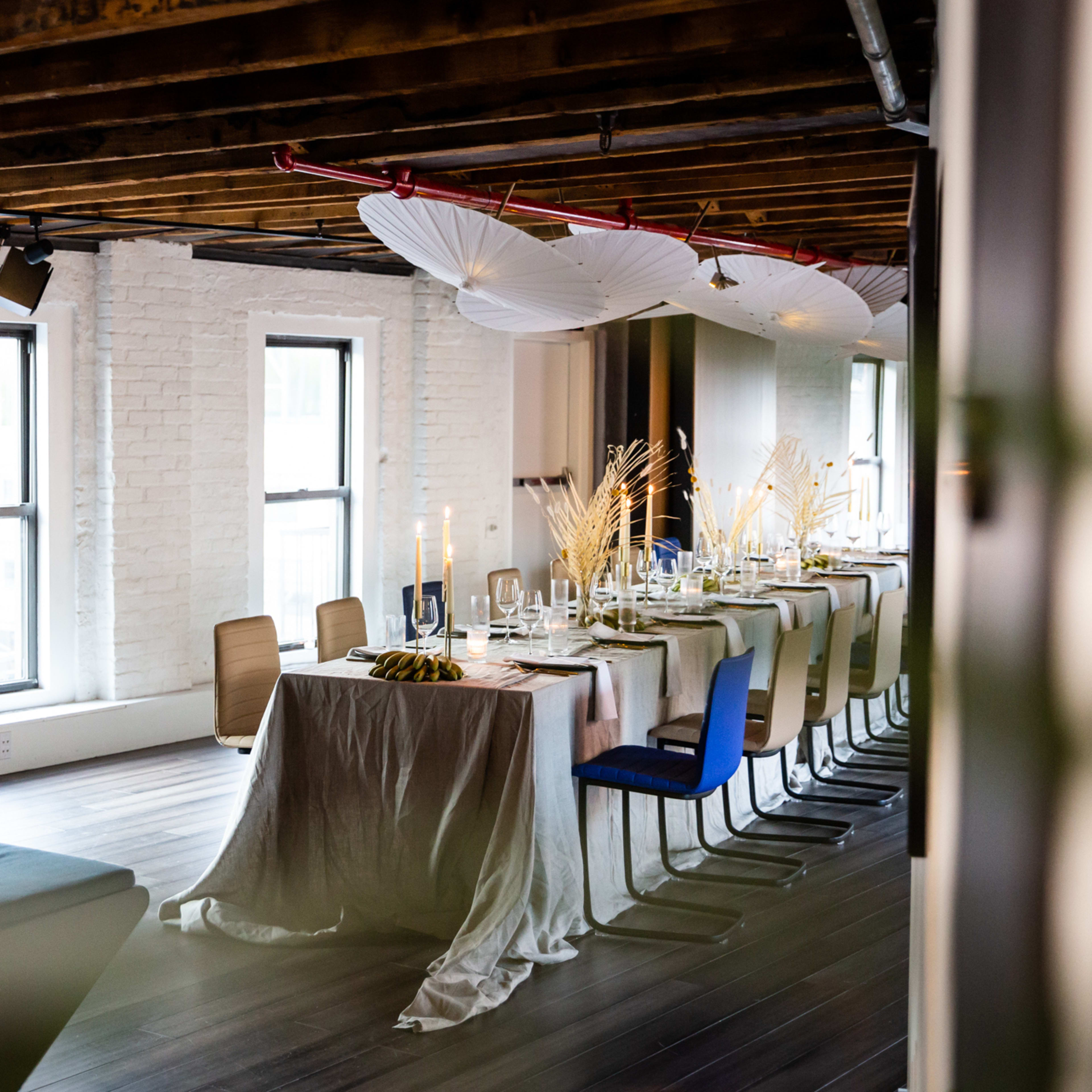 A long dining table is set with elegant tableware and decorated with white umbrellas and dried flowers in a modern interior space.