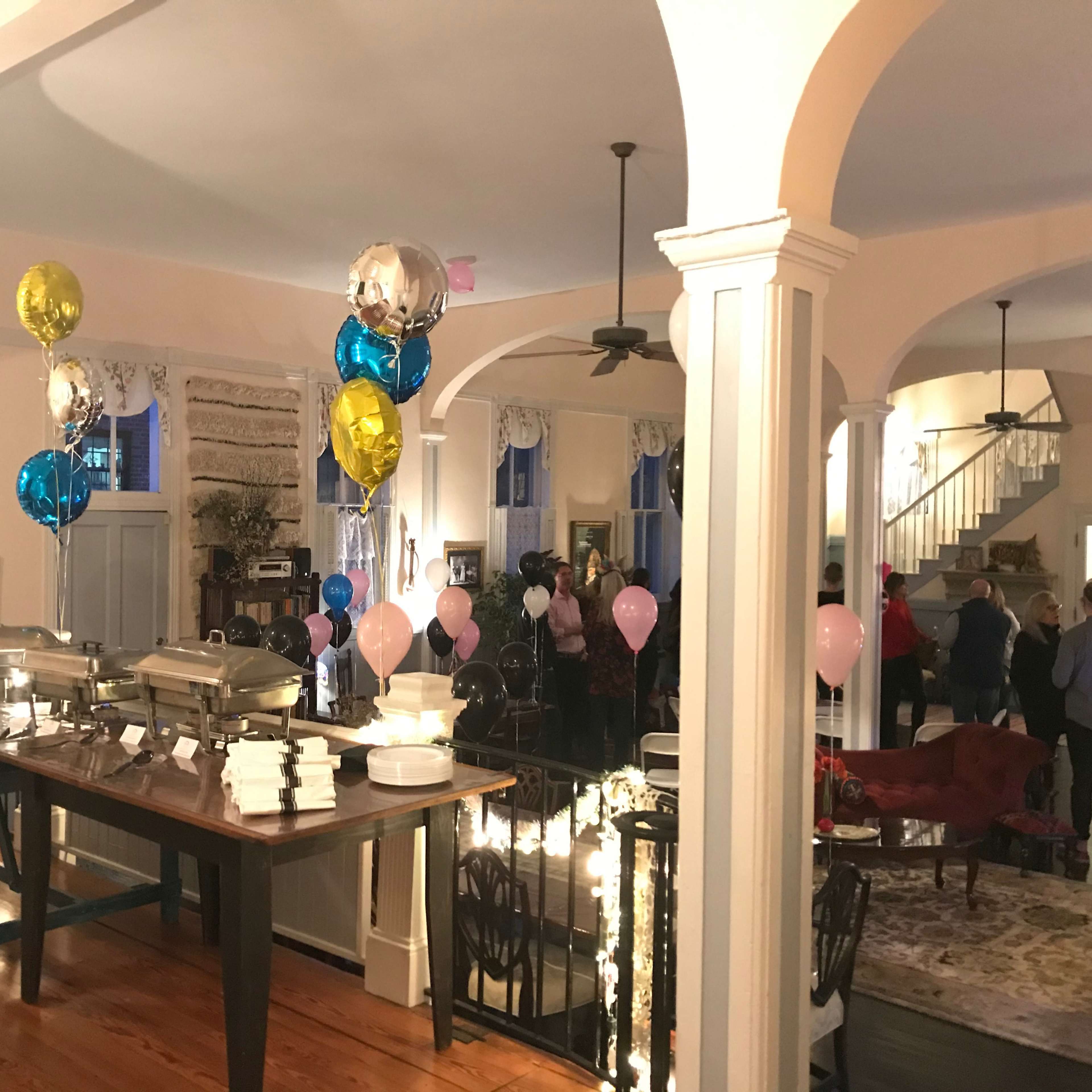 A spacious indoor party setting with a table displaying food trays, colorful balloons, and guests mingling in the background.