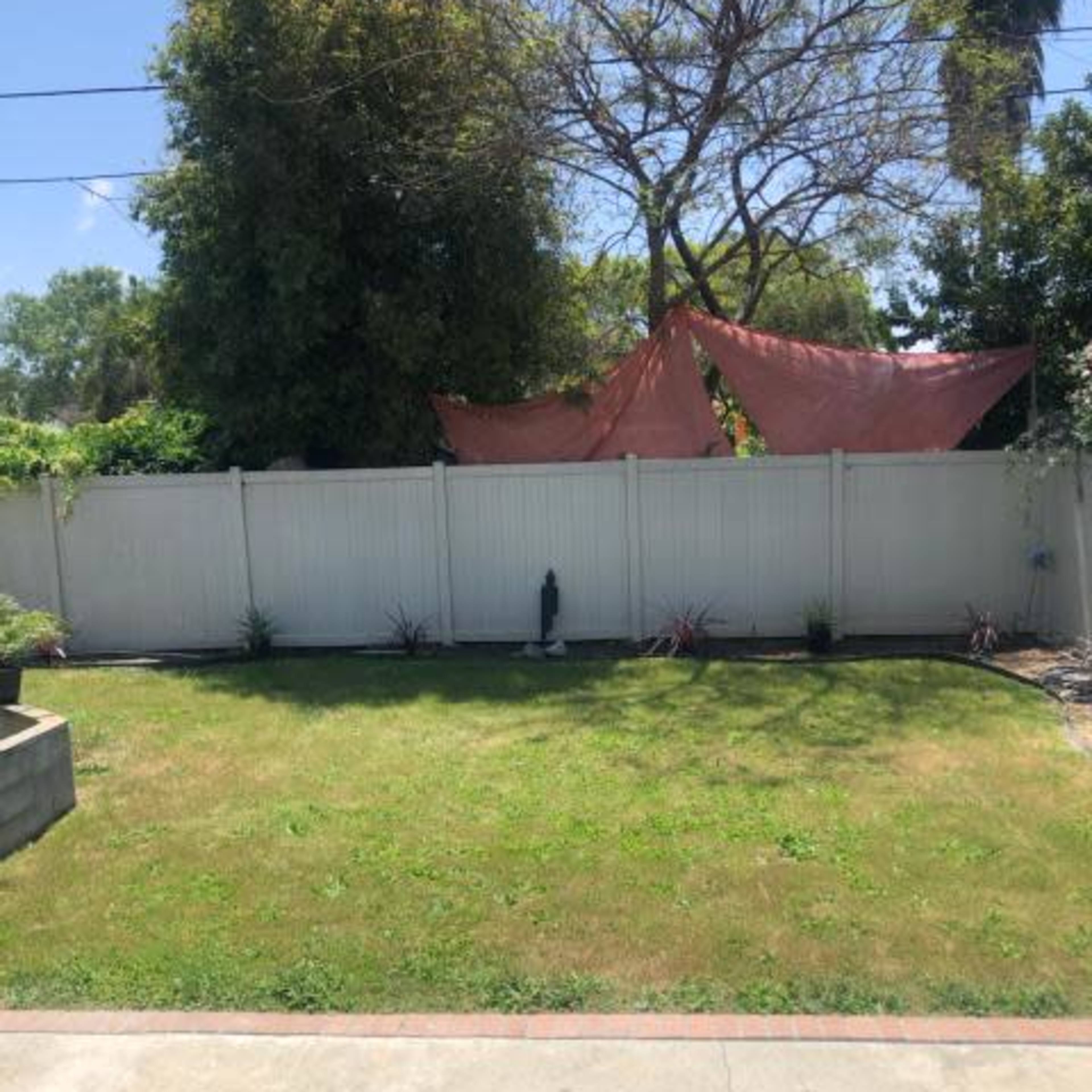 A fenced backyard features a patch of grass, a statue in the center, and some plants along the perimeter, with a large tree and a structure covered by a fabric sheet in the background.