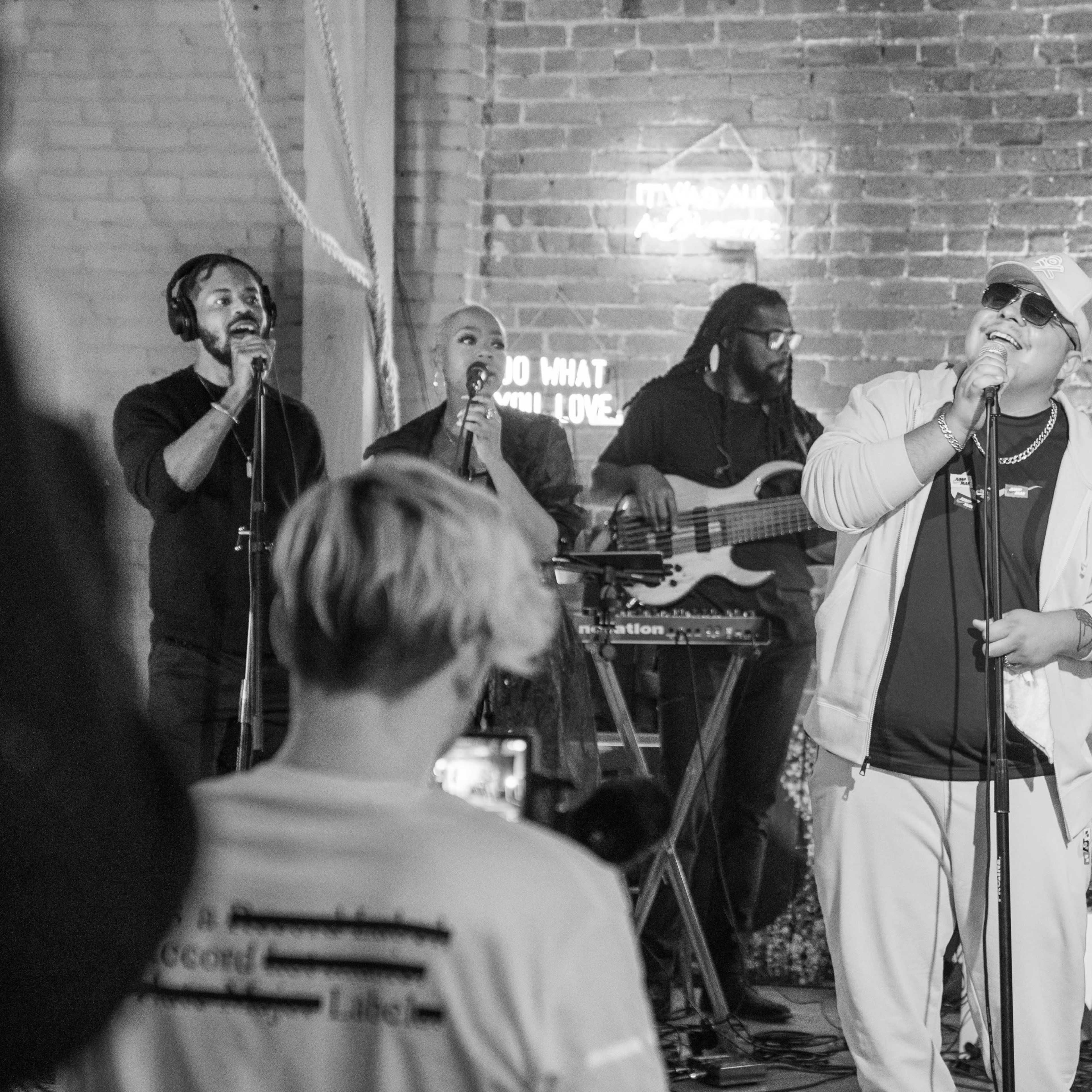 A group of musicians performs on stage with a brick backdrop, featuring a singer in a white outfit and sunglasses, accompanied by backup vocalists and instrumentalists.