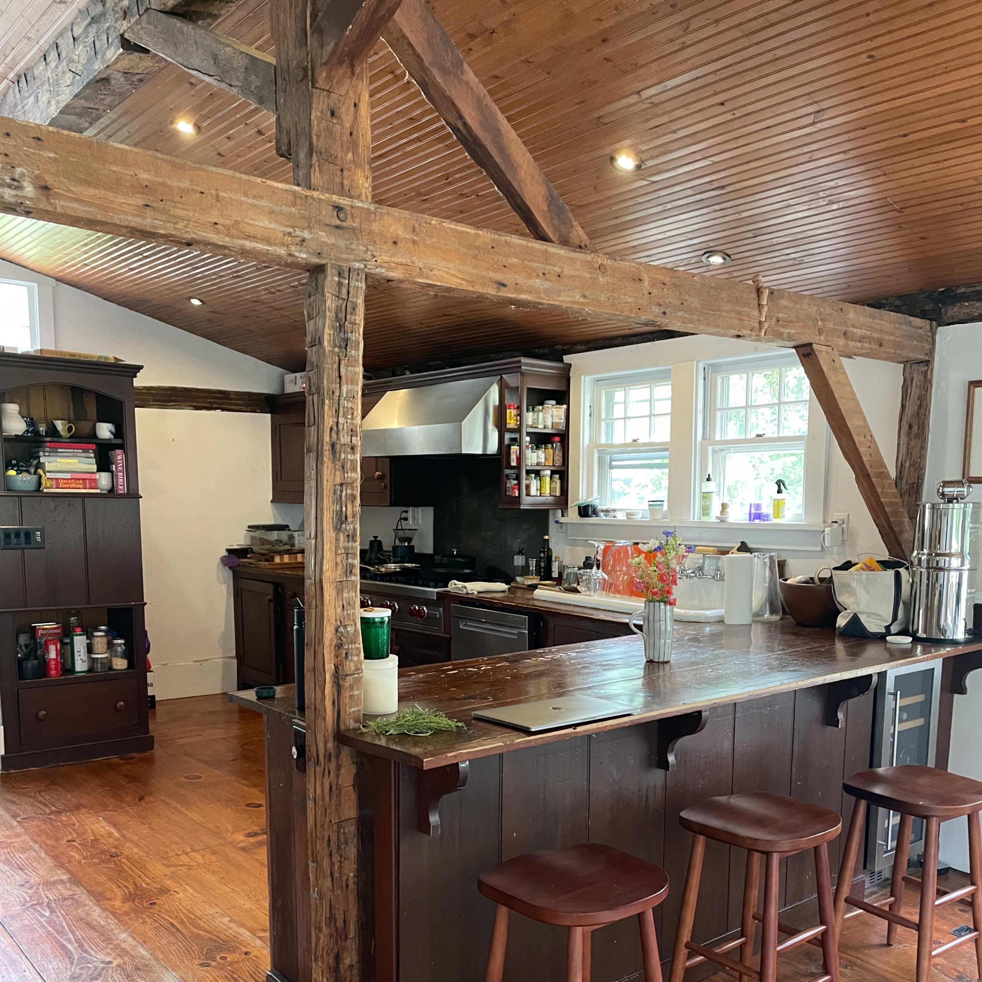 A rustic kitchen with wooden beams, a central island with three stools, and cabinets lining the walls.