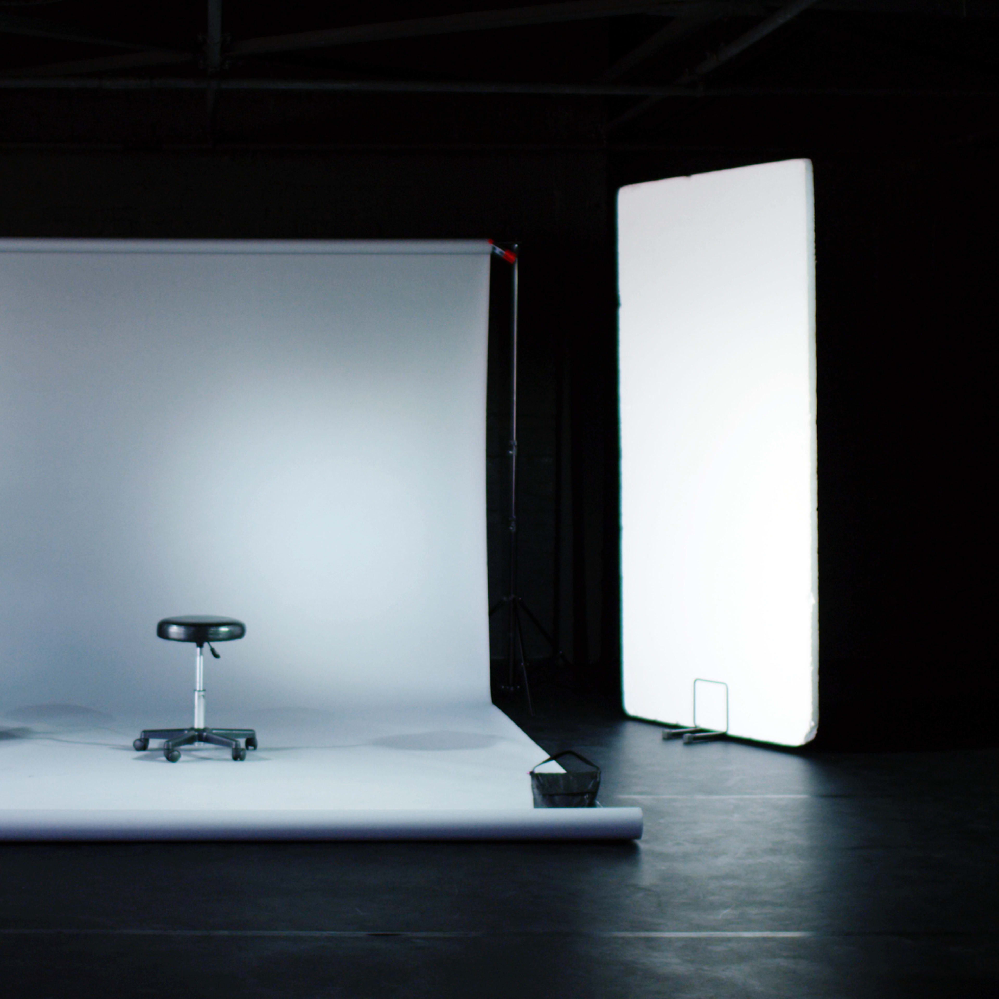 An empty photo studio setup with a backdrop, a stool in the center, a ladder, a fan, and a softbox on the right.