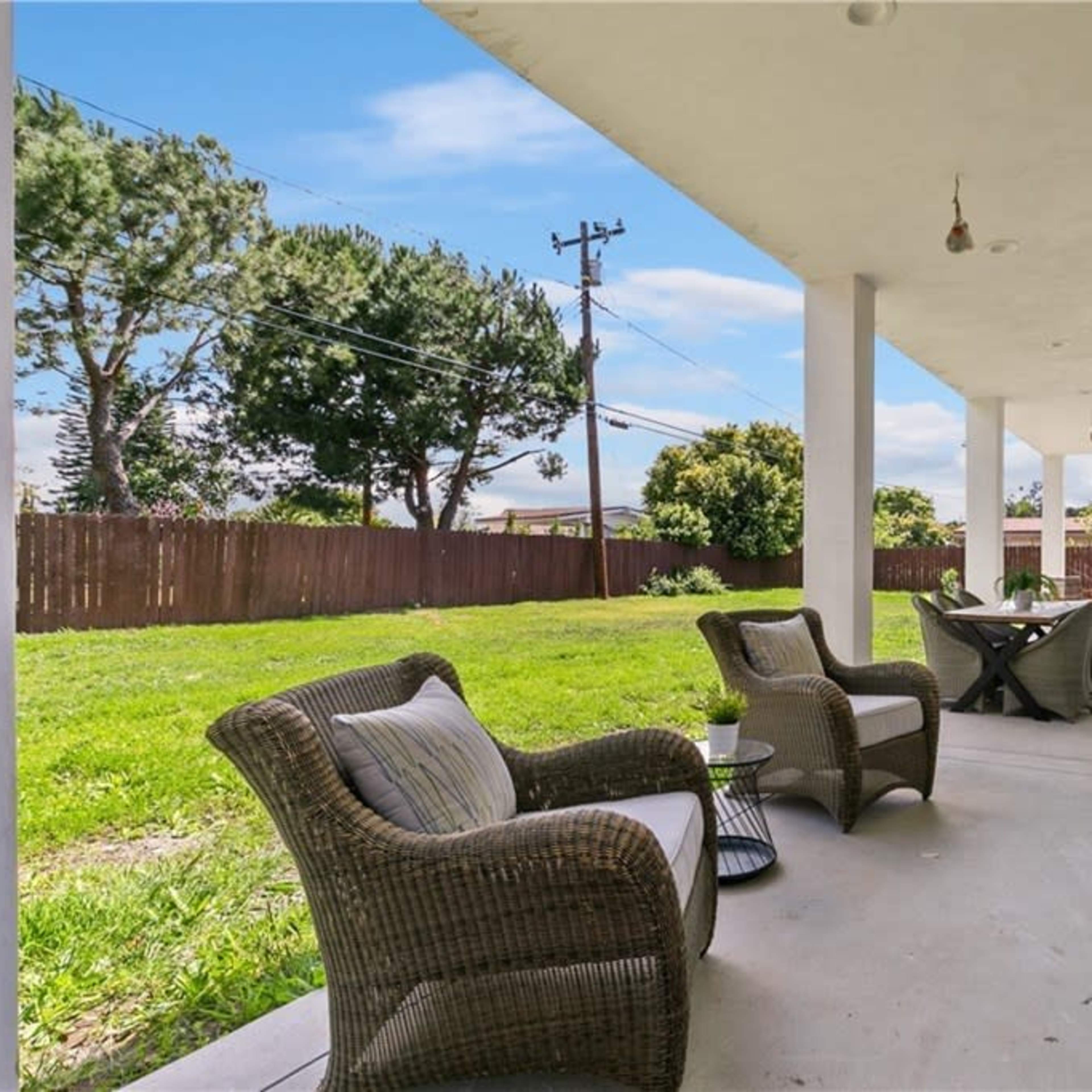 A covered patio features two chairs and a table, overlooking a grassy yard with trees in the background.