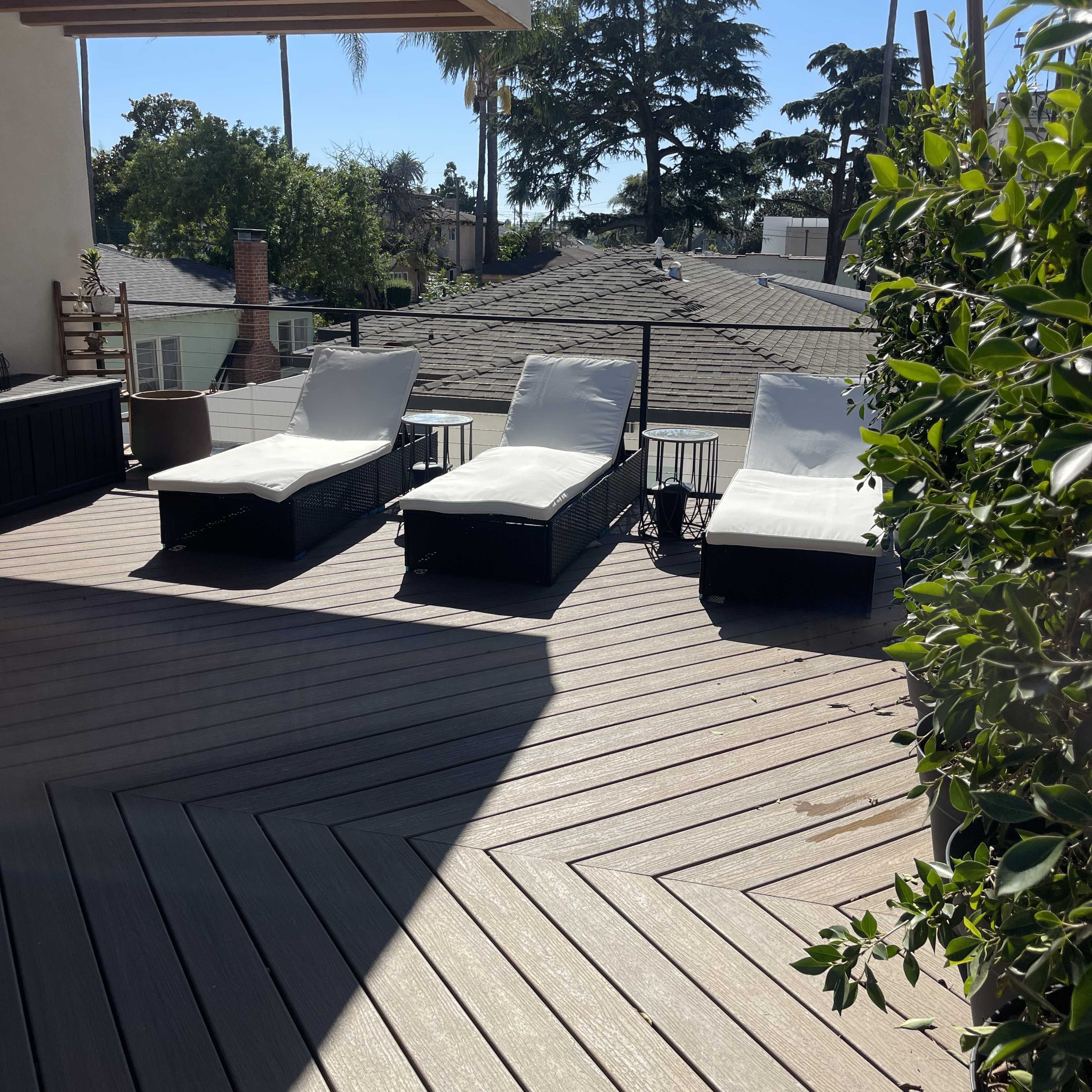 Three white lounge chairs on a wooden deck under clear blue skies, surrounded by greenery and rooftops.