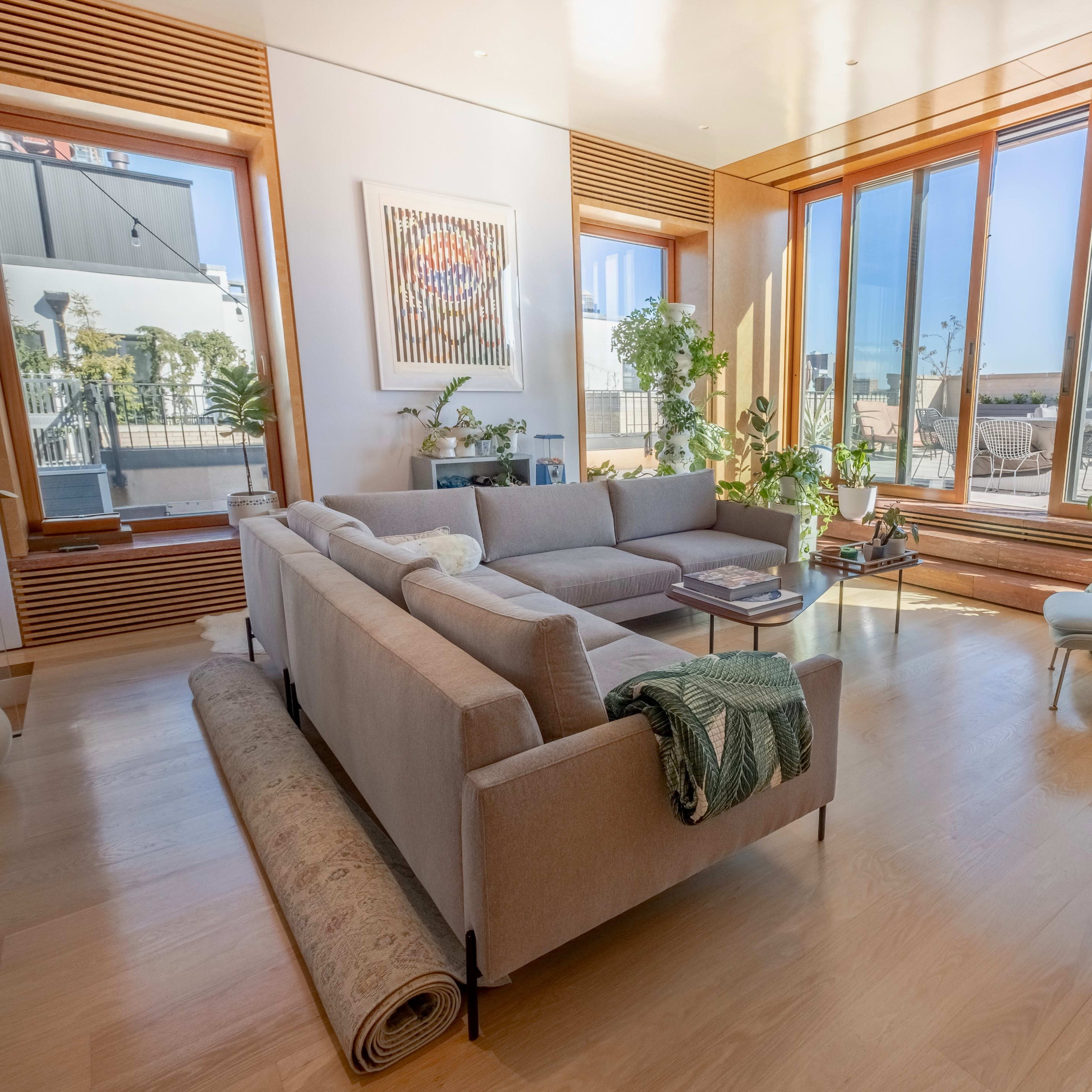 A bright living room features a large sofa, potted plants, and expansive windows with a view of the city.