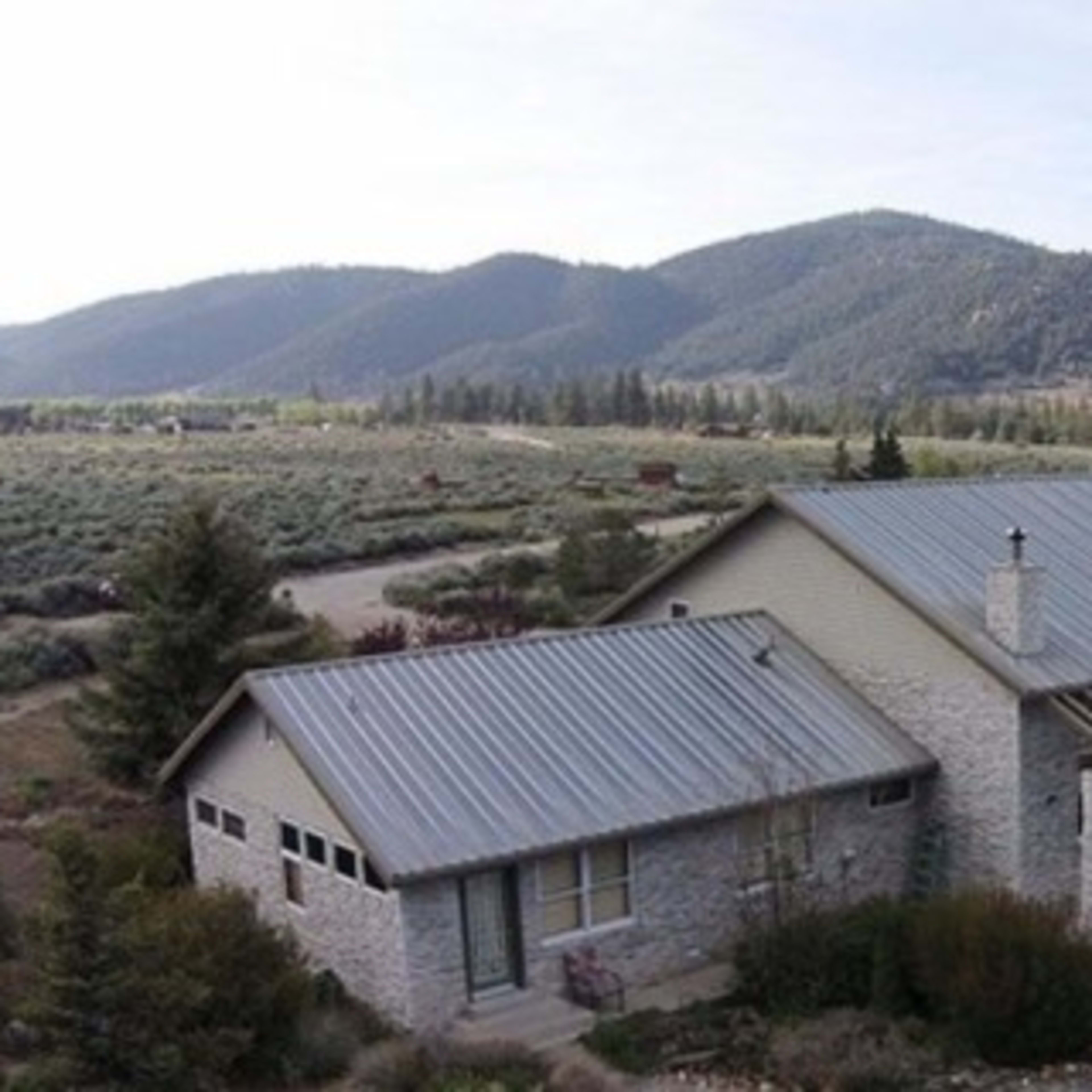 A large, single-story house with a metal roof situated near rolling hills and mountains in a rural area.