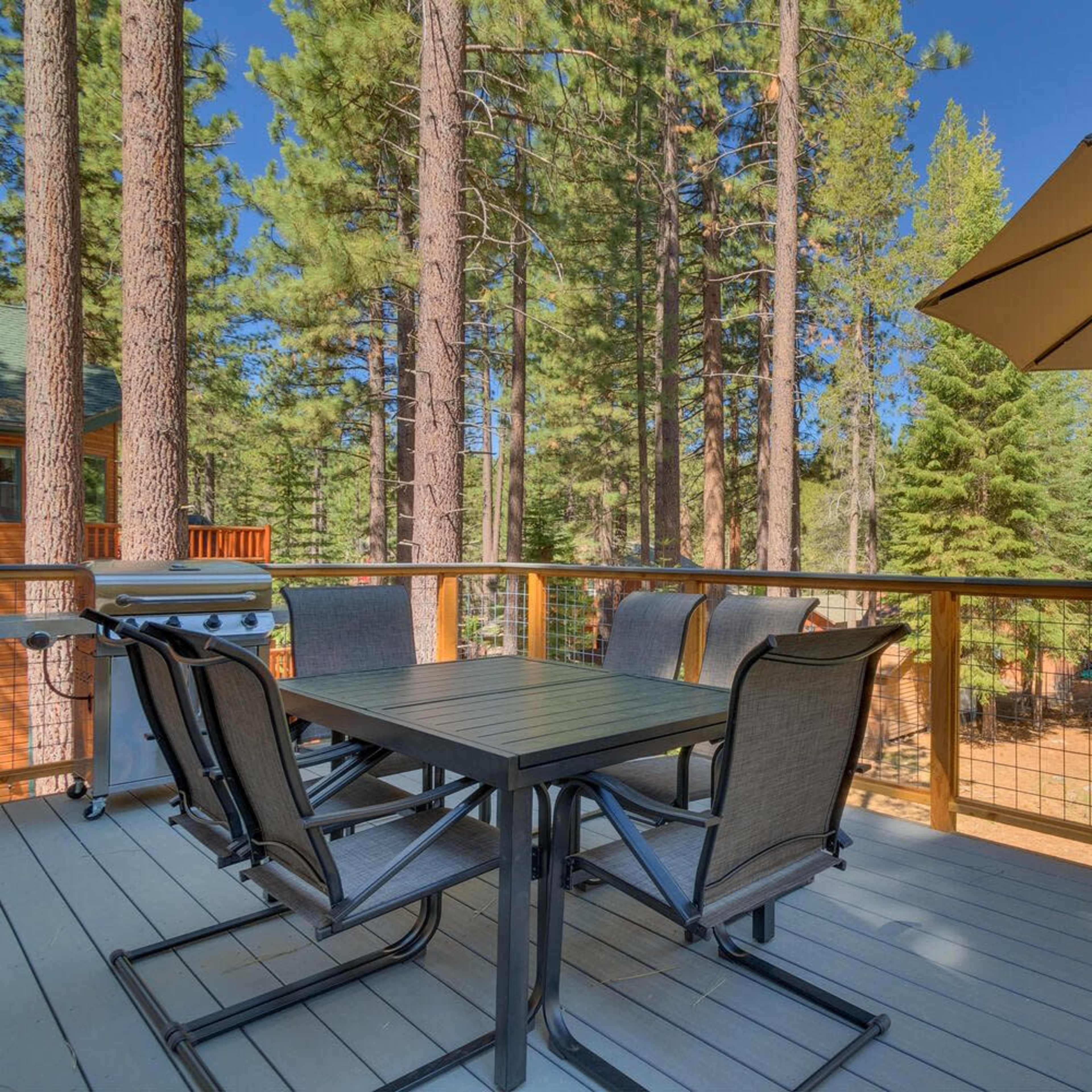 A wooden deck features a dining table with six chairs and an umbrella, surrounded by tall trees in a forested area.