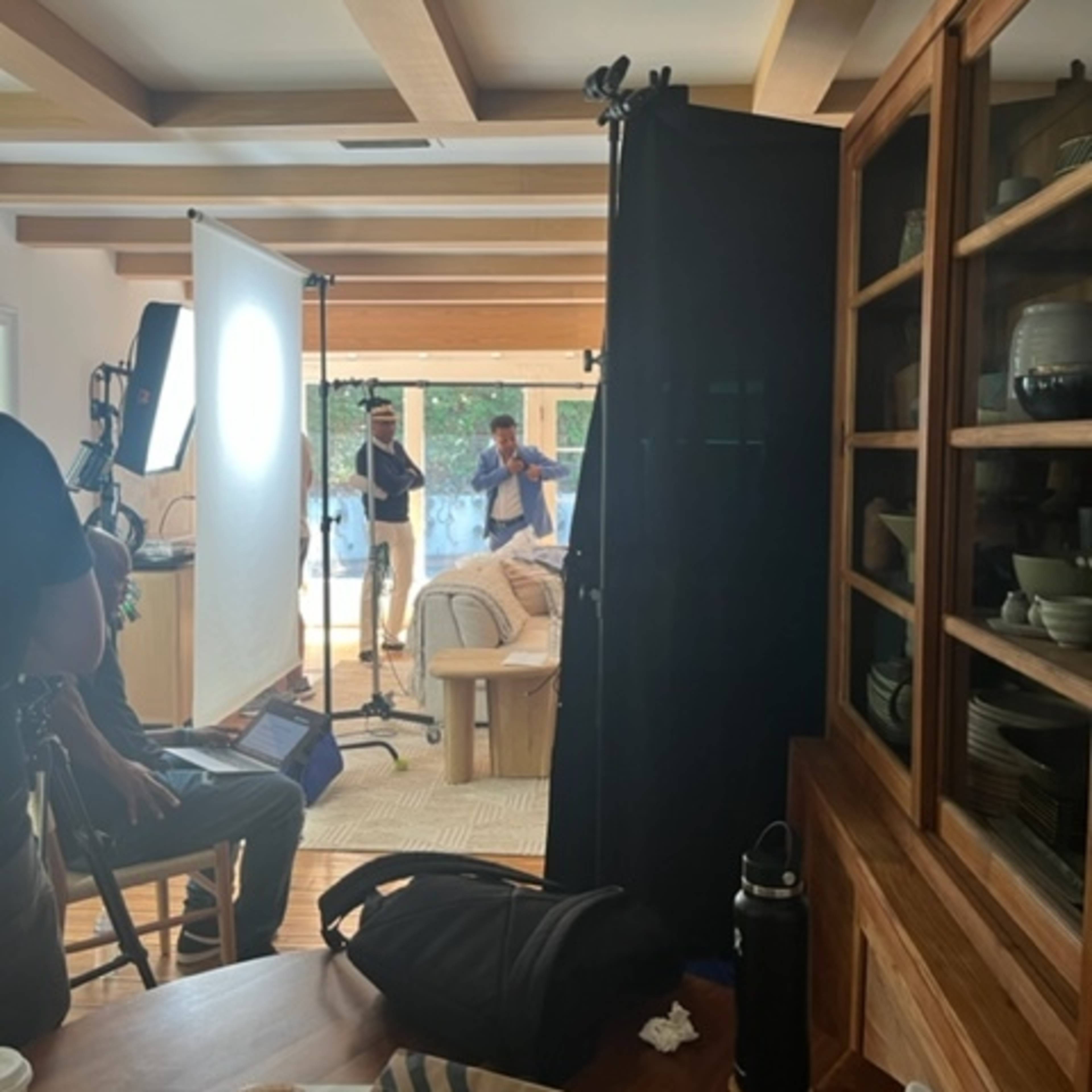 A film crew is set up inside a room, with a camera aimed at two people on a couch, and studio lighting positioned nearby.