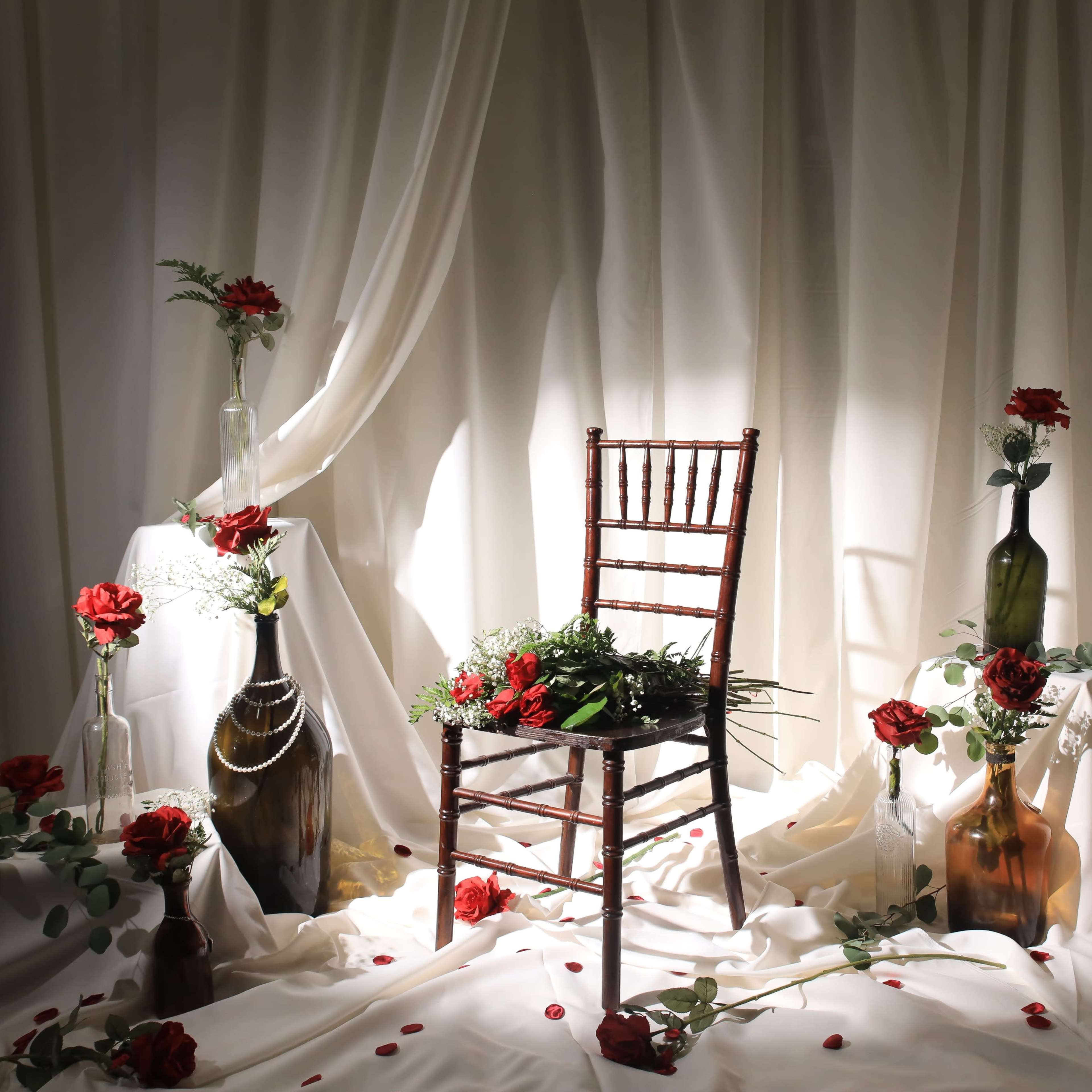 A wooden chair surrounded by vases of flowers and draped fabric creates a simple, elegant arrangement.