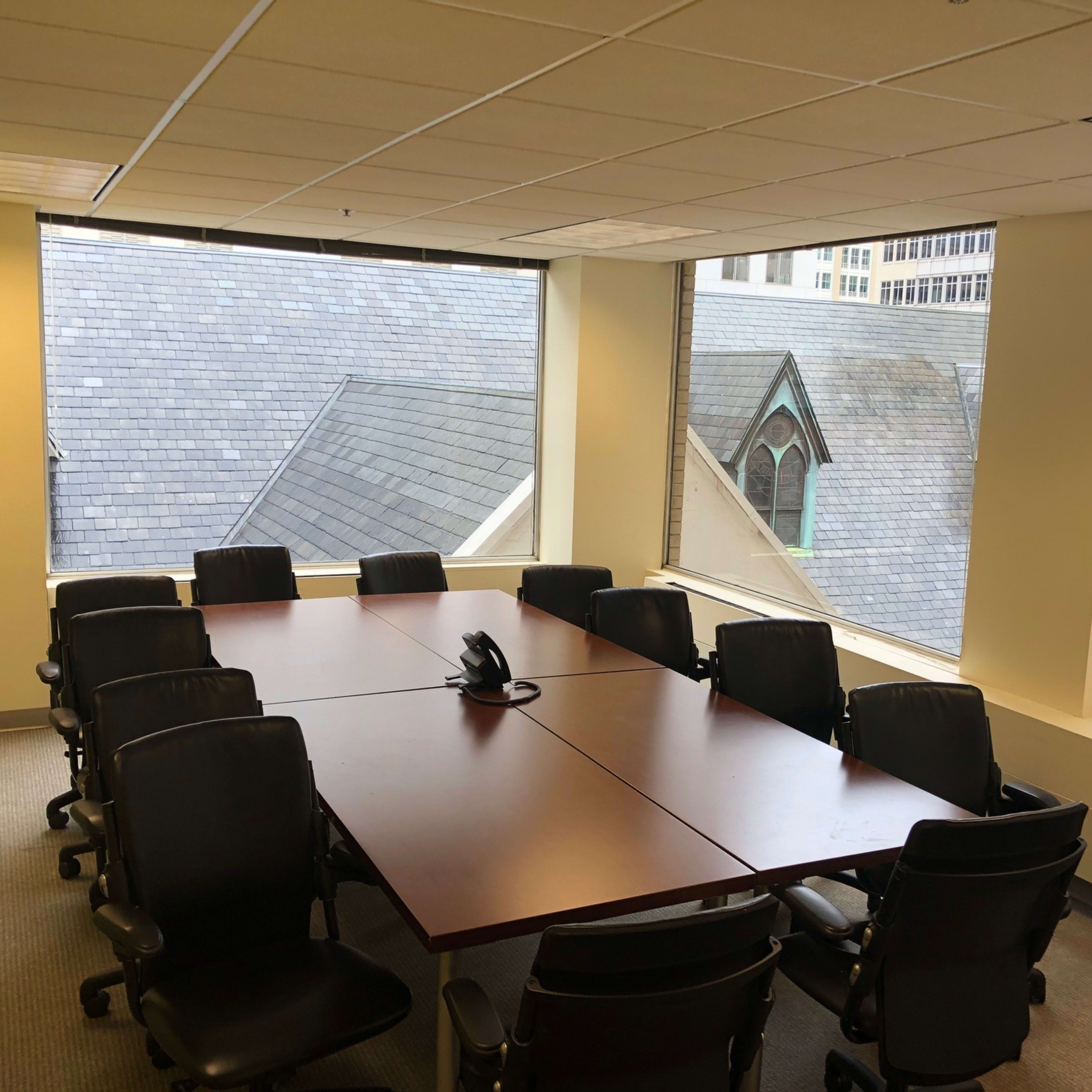 A conference room with a rectangular wooden table surrounded by black chairs and large windows overlooking rooftops.
