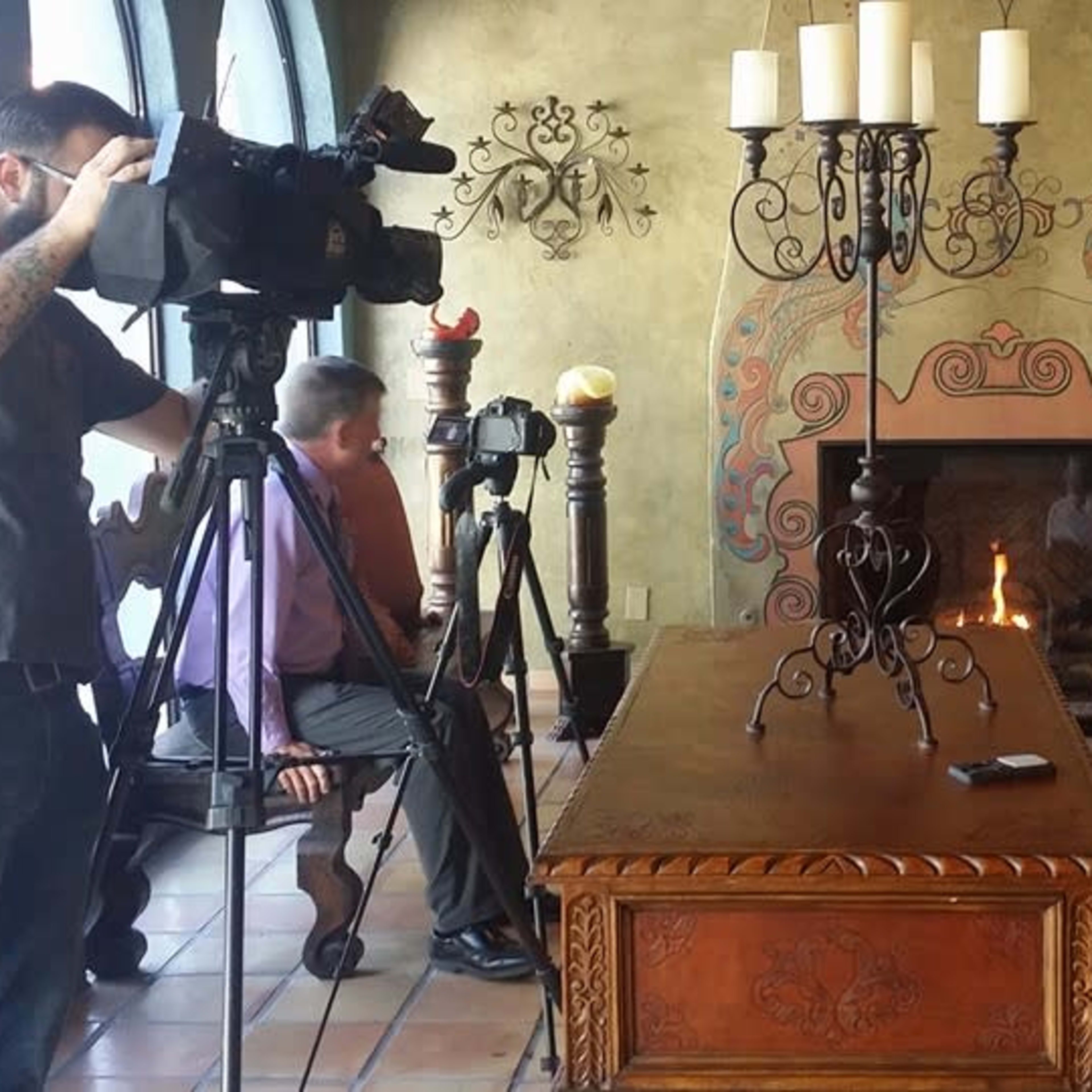 A woman sits on a couch in a decorative room while two men operate cameras nearby.