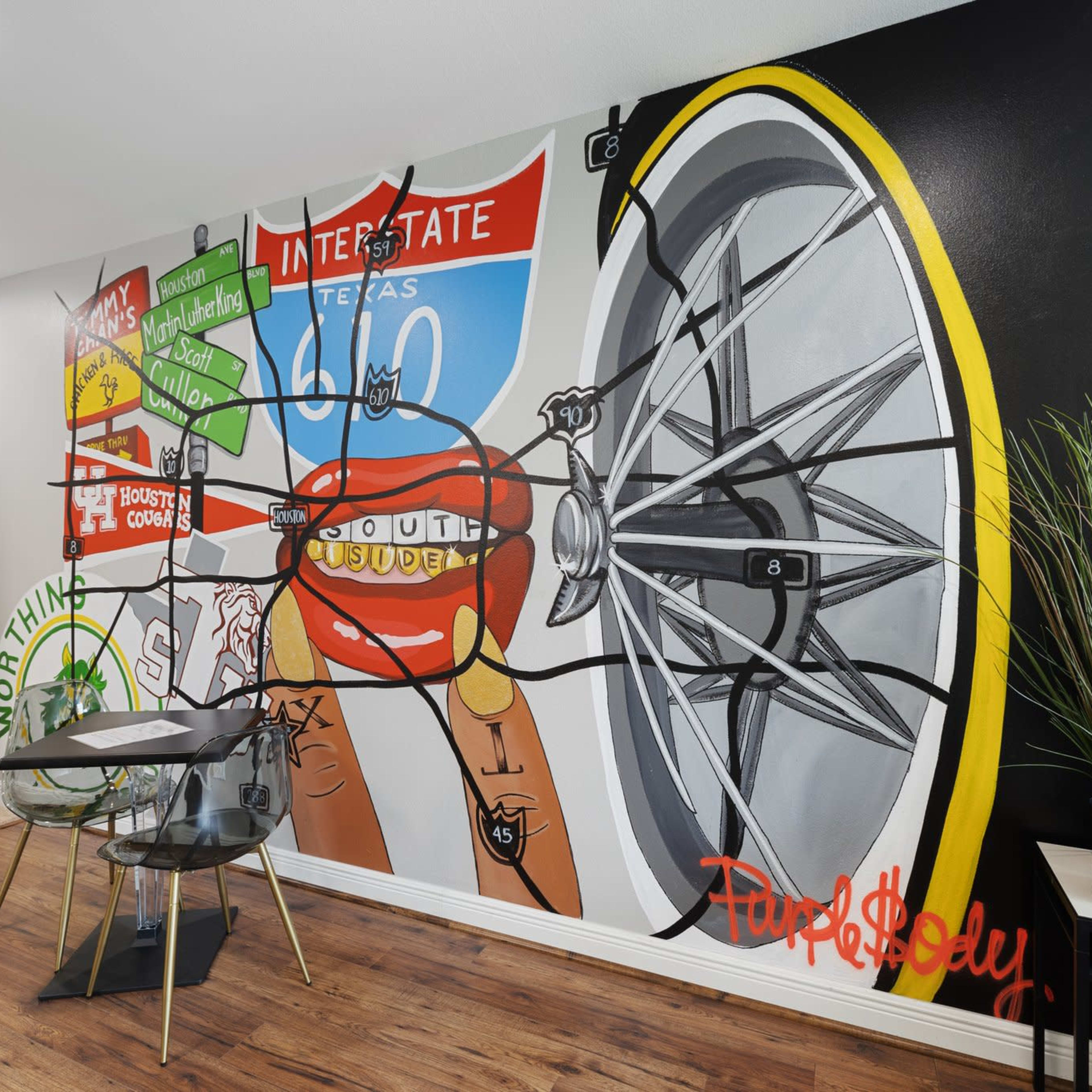 A vibrant mural on a wall featuring a bicycle wheel, a Texas highway sign, and playful graphics along with a small dining table and chairs set nearby.