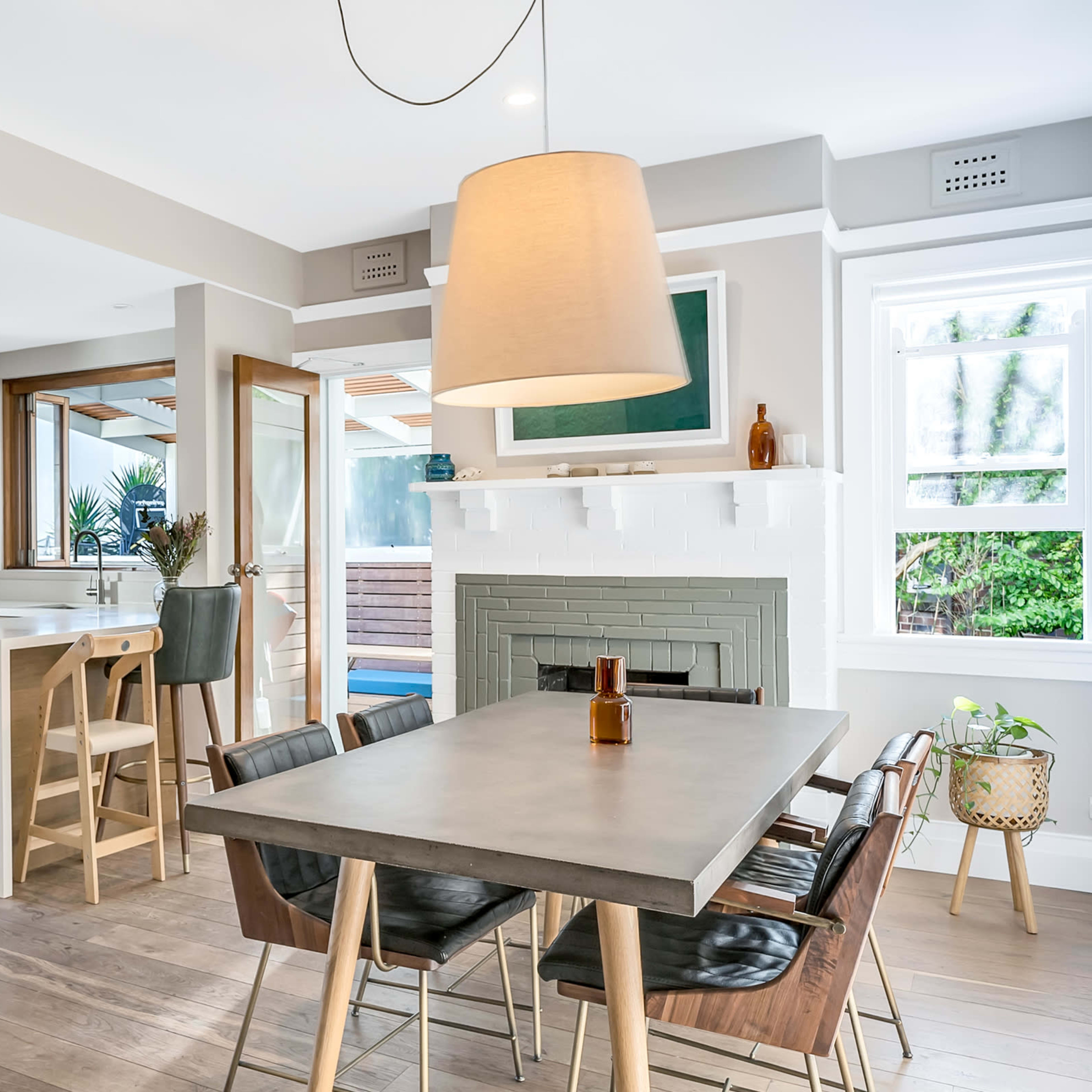 A modern kitchen and dining area with a concrete dining table, wooden chairs, and a green fireplace, featuring large windows that provide natural light.