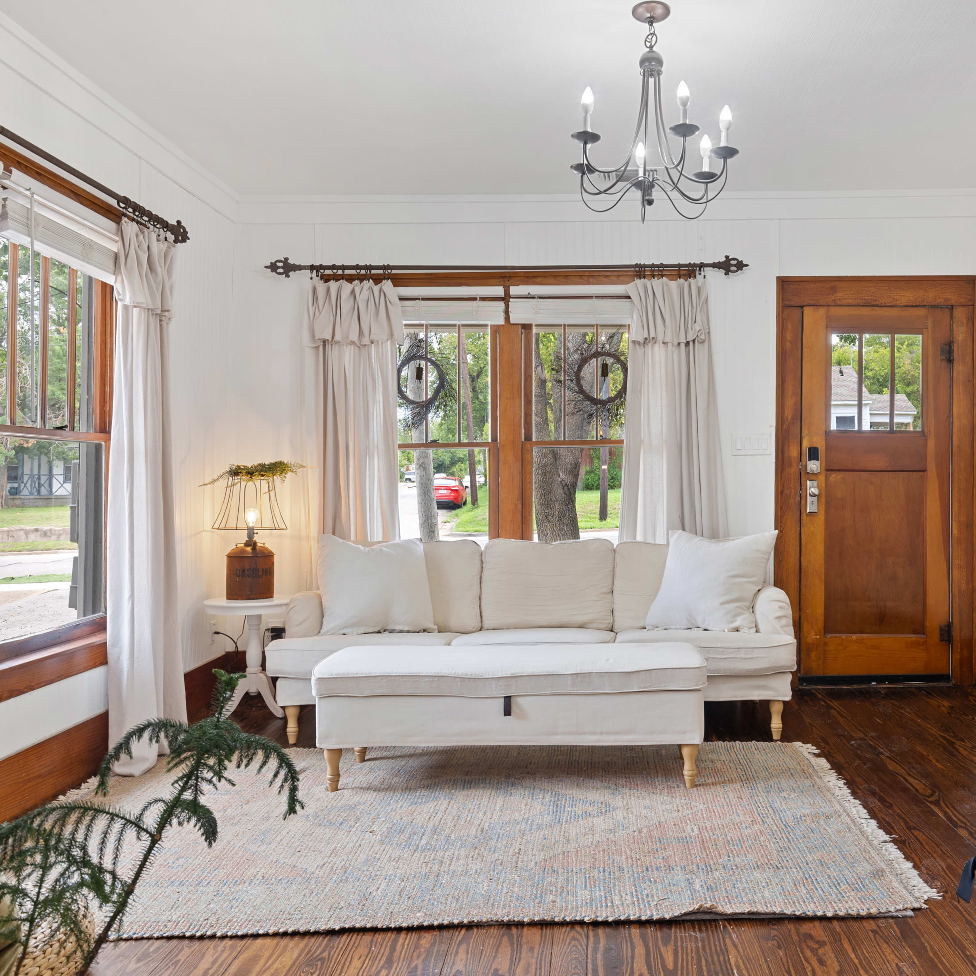 The living room features a white sofa, a wooden door, and a chandelier, along with large windows framed by curtains.