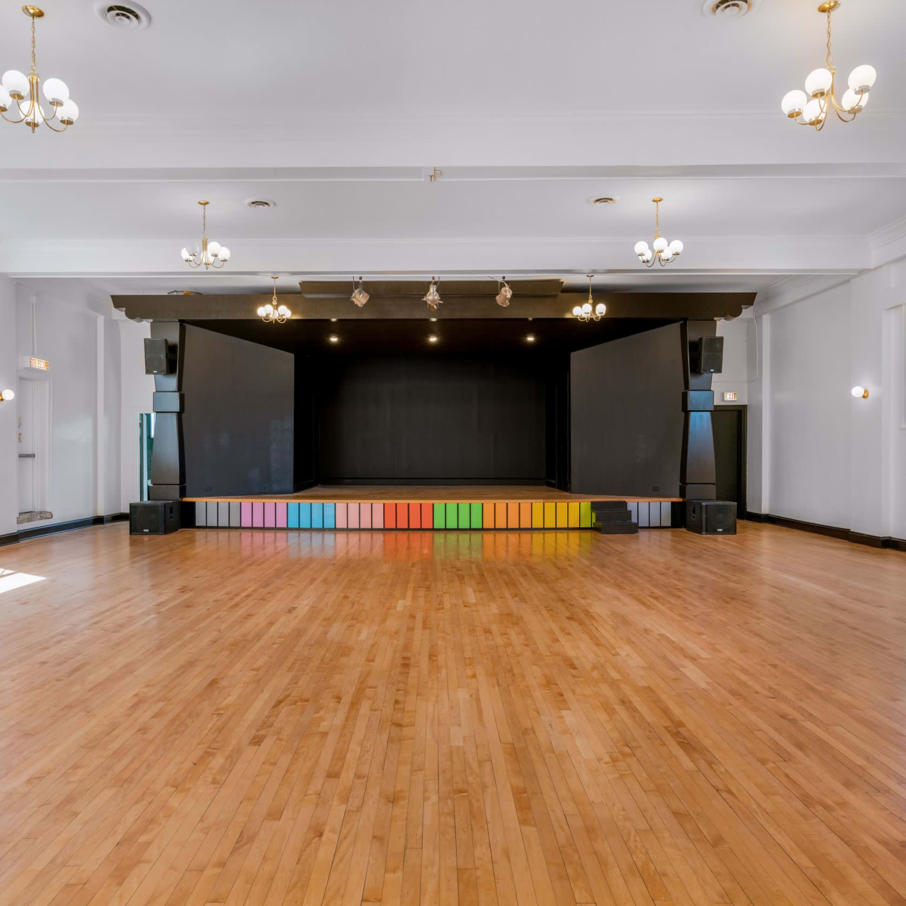 A spacious, empty hall with polished wooden floors, a stage at the front featuring colorful steps, and bright lighting from overhead chandeliers.