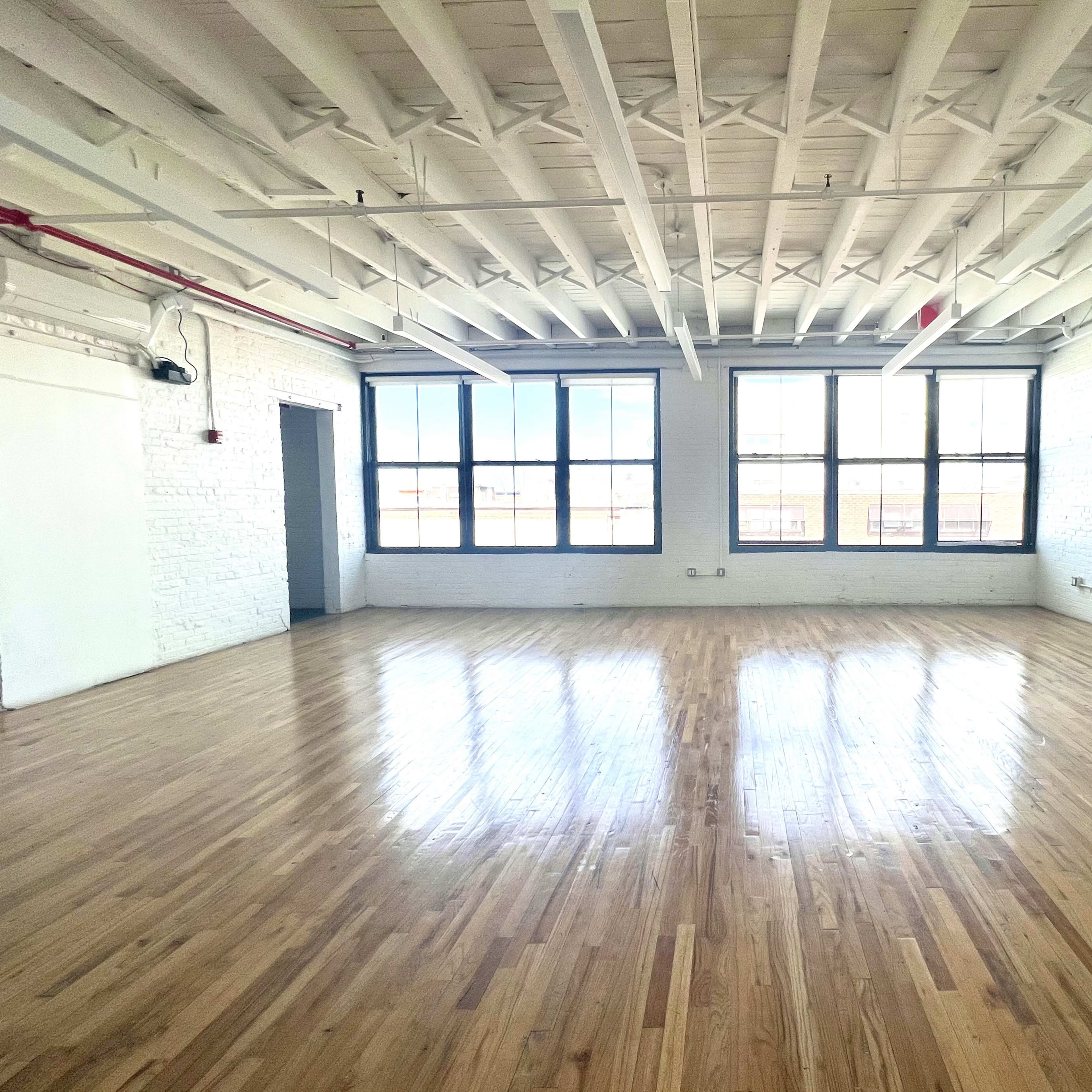 A spacious, empty room with hardwood floors and large windows along one wall, illuminated by natural light.